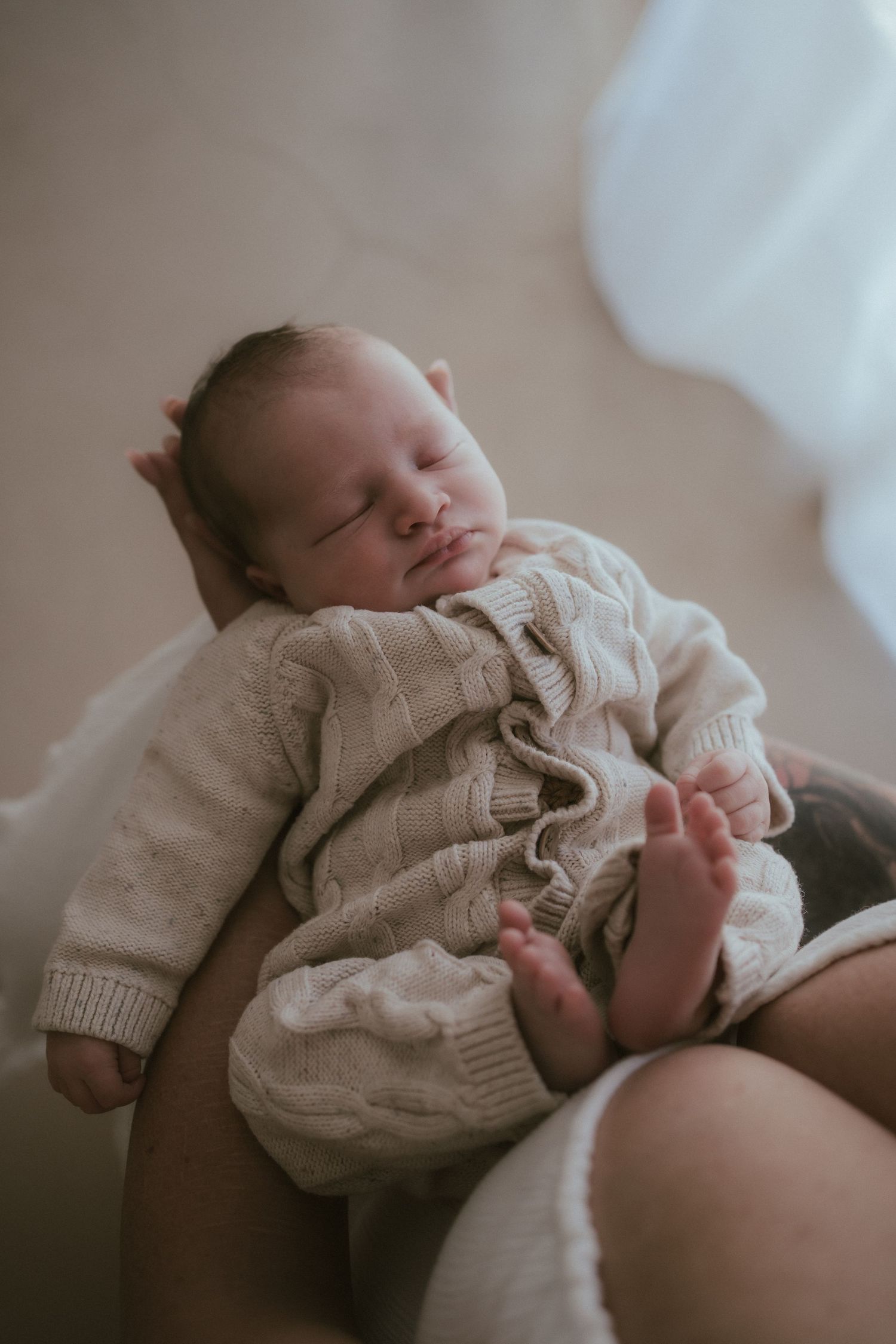 A peaceful sleeping newborn wearing a cozy knit cream sweater lies on soft bedding in natural light.