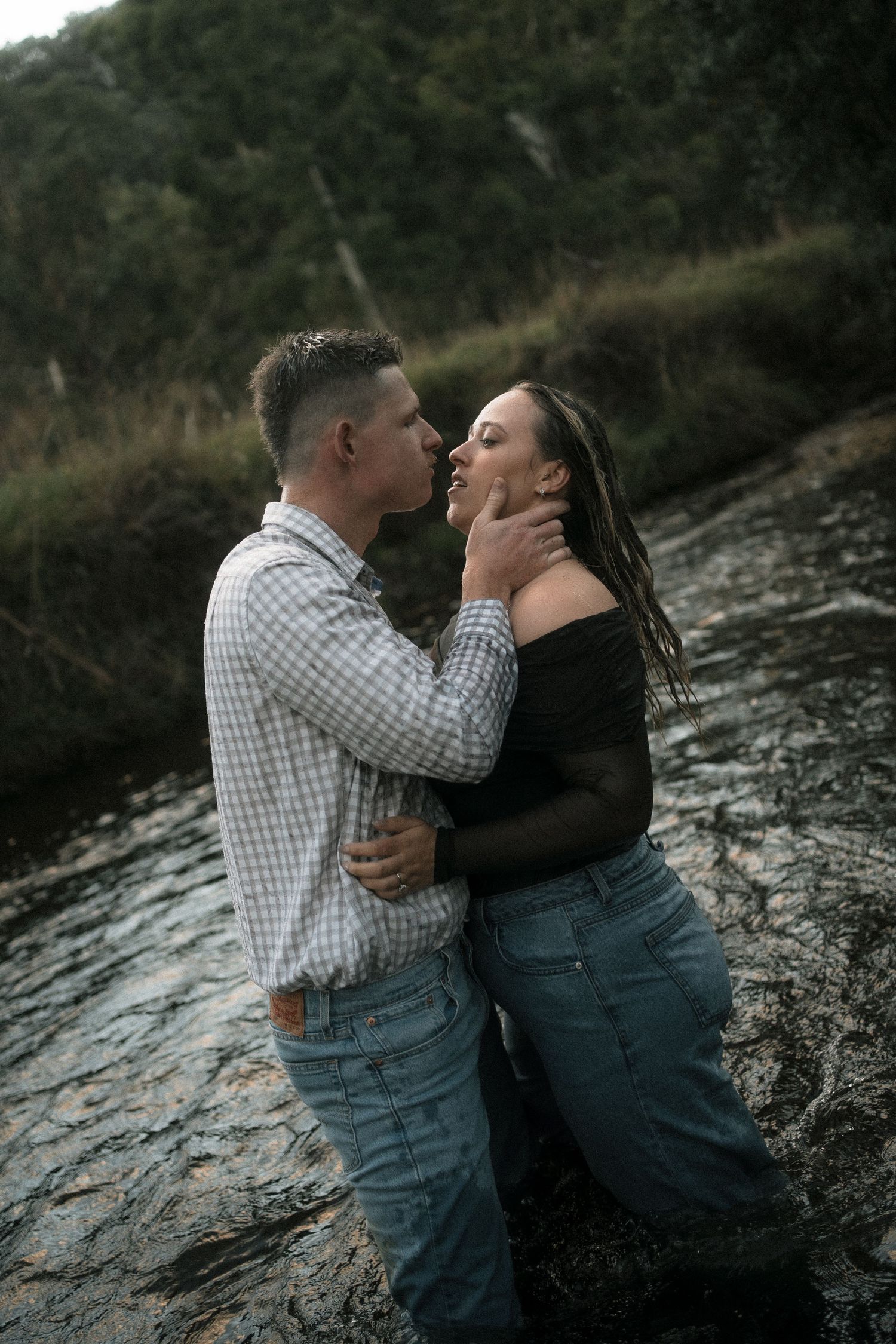 Couple sharing a romantic moment on wet rocks in nature, man in plaid shirt and woman in black top and jeans.