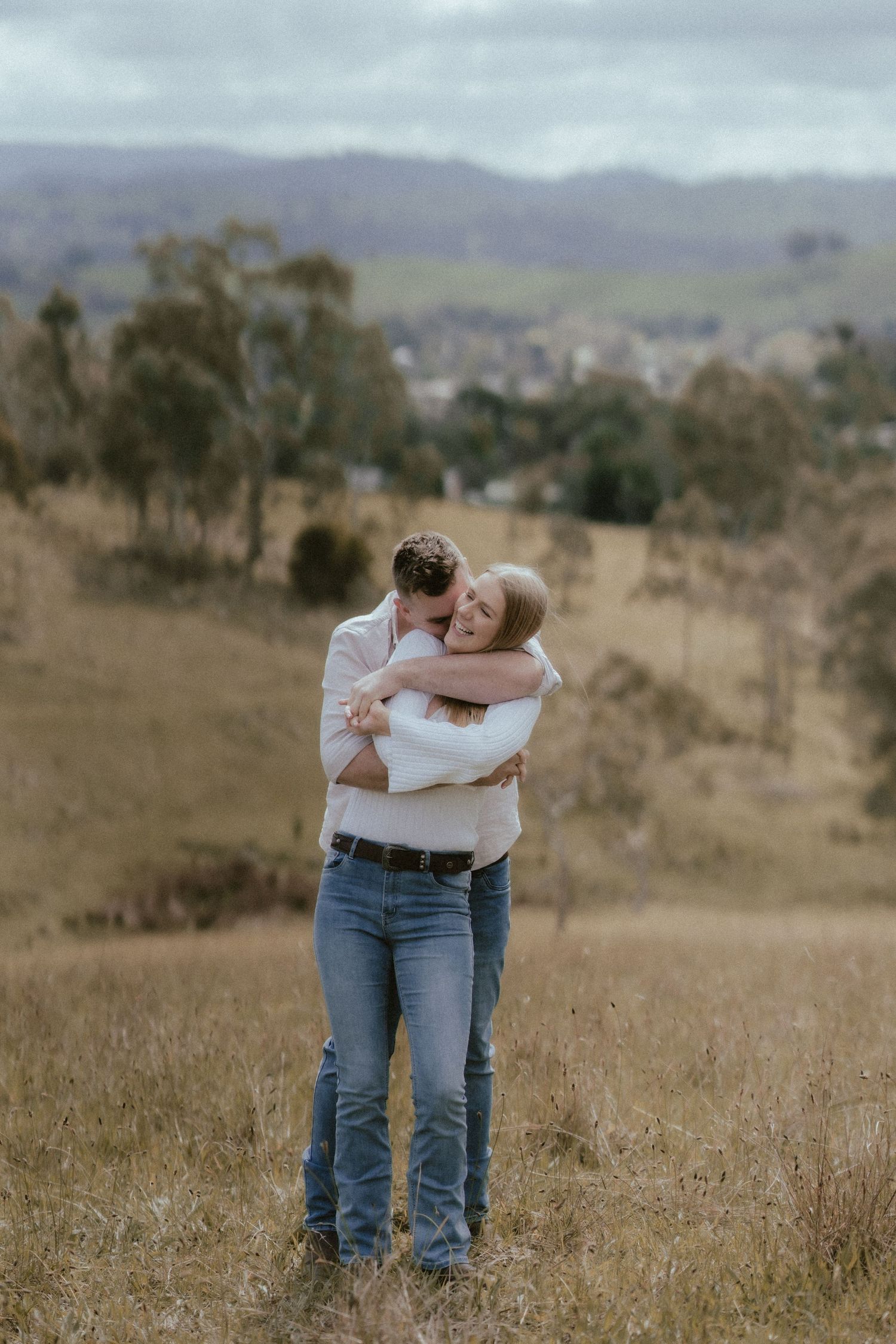 A couple embraces in a tender moment on a hilltop overlooking a distant town with mountains and eucalyptus trees in view.