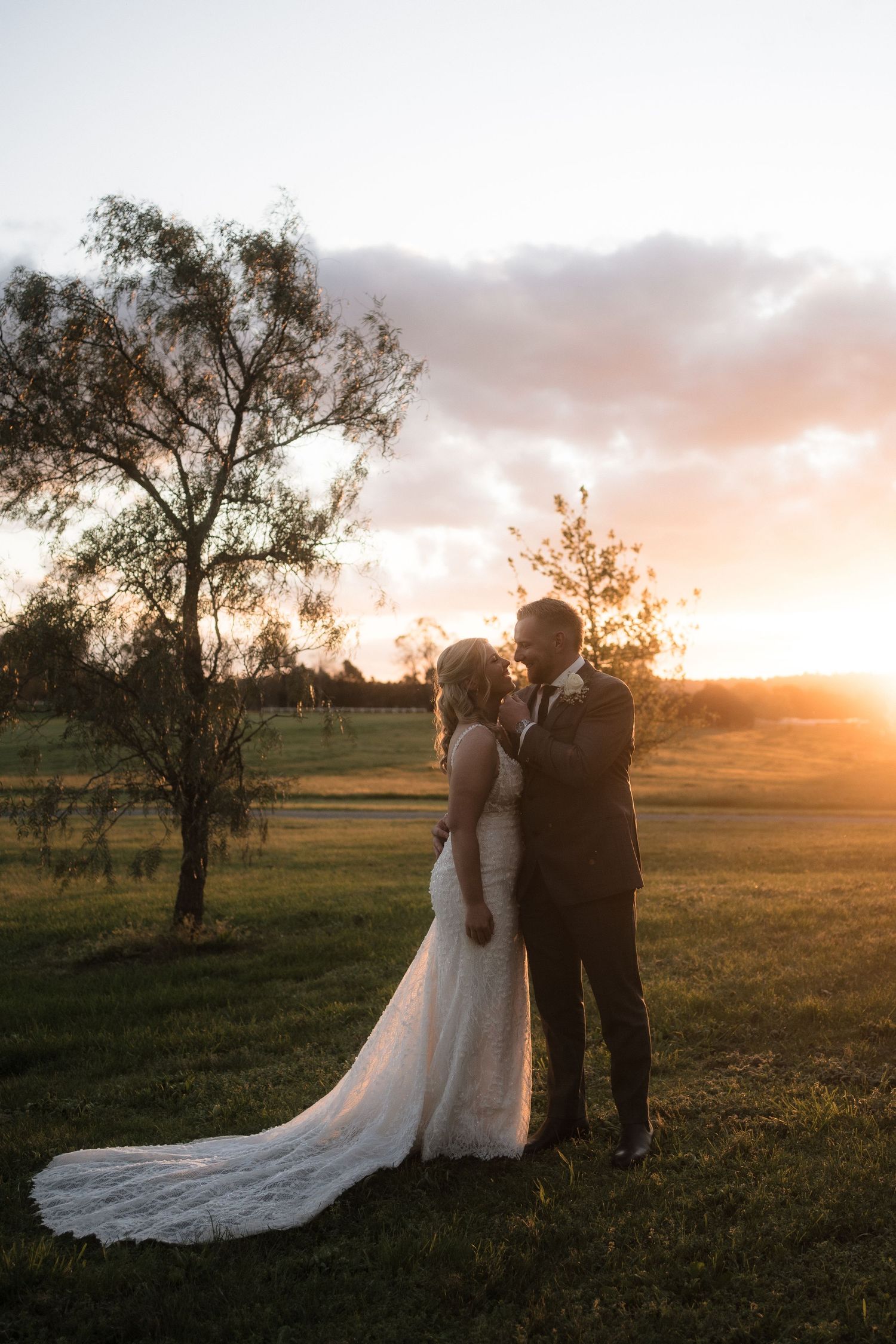 A couple stands together in a field at sunset as golden light illuminates their silhouettes against a dreamy rural landscape.