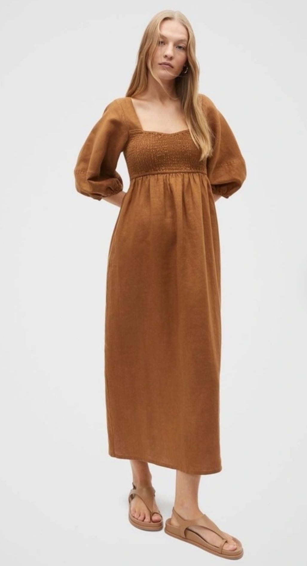 Long brown empire-waist dress with puff sleeves and smocked bodice, paired with tan leather sandals against white background.