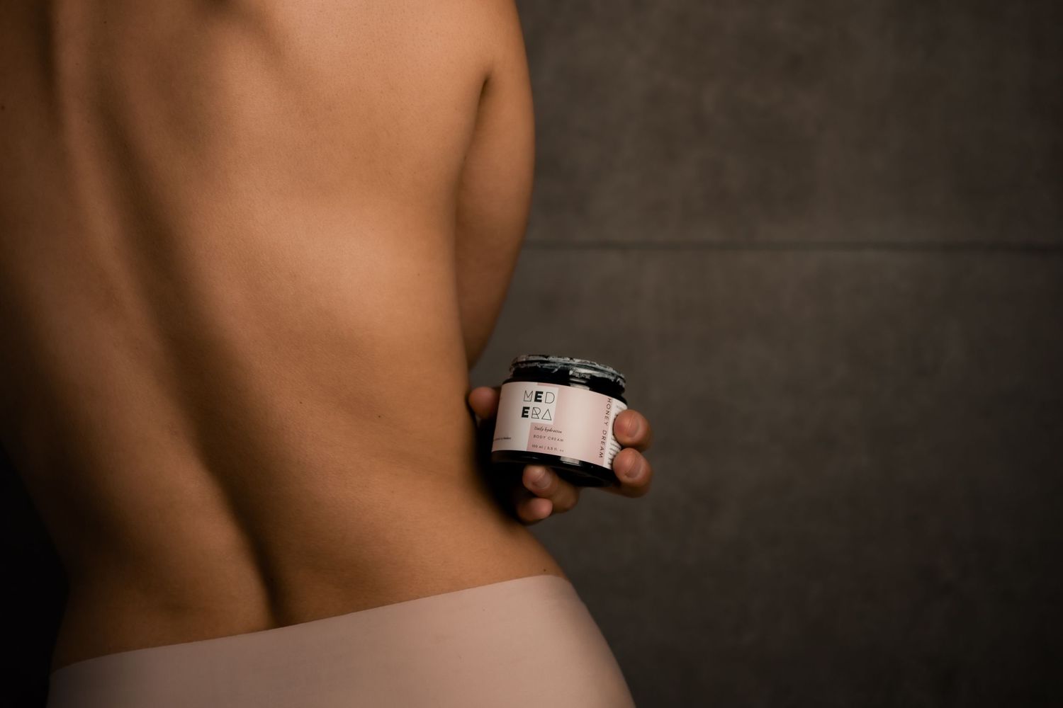 A bare-skinned torso holding a skincare product jar against a dark background.