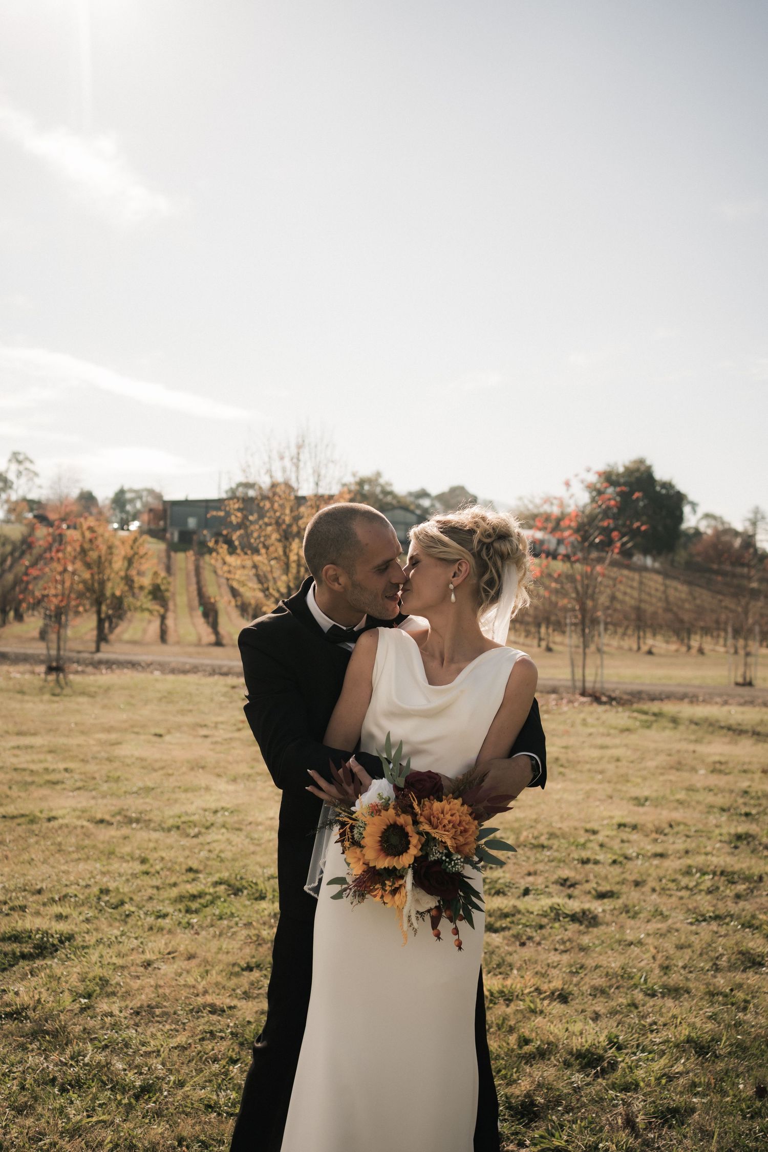 A couple shares an intimate moment in a rustic field setting with fall foliage and sunflowers in the bridal bouquet.