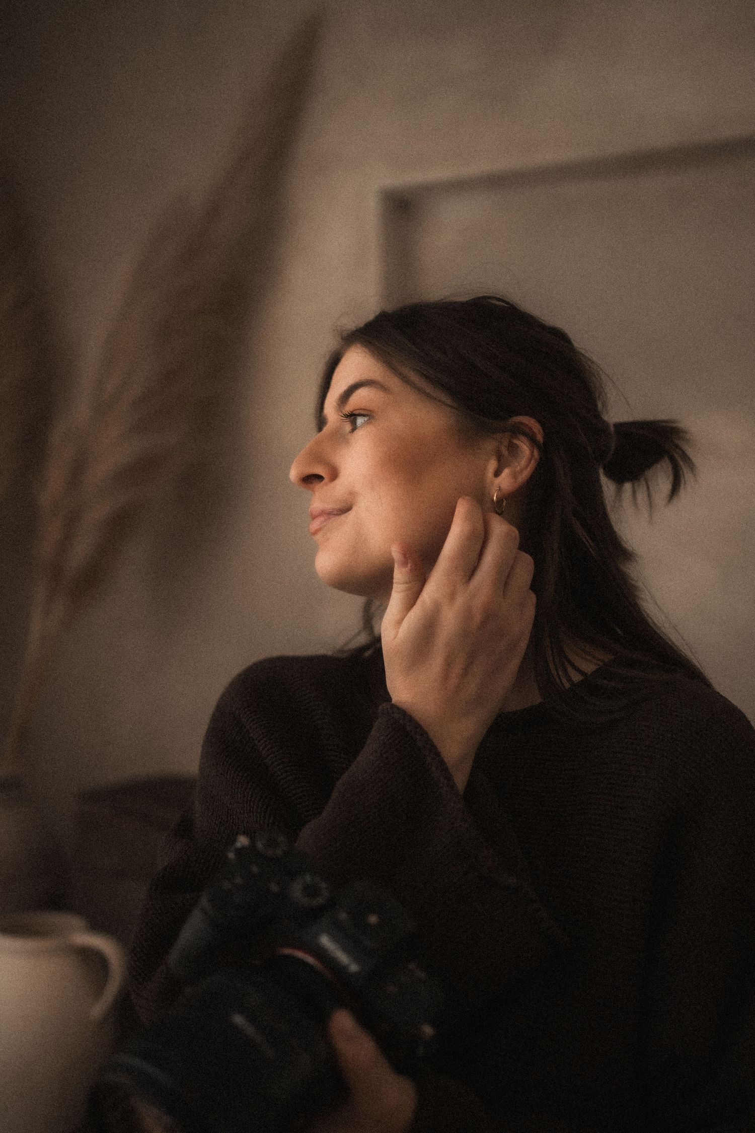 Woman with dark hair in a bun gazes out a window, hand resting on her cheek, holding a camera in a softly lit, neutral-toned room.