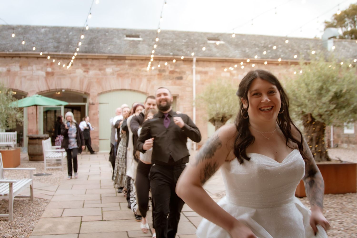 Bride in white gown leading wedding party across a courtyard with festive string lights overhead.