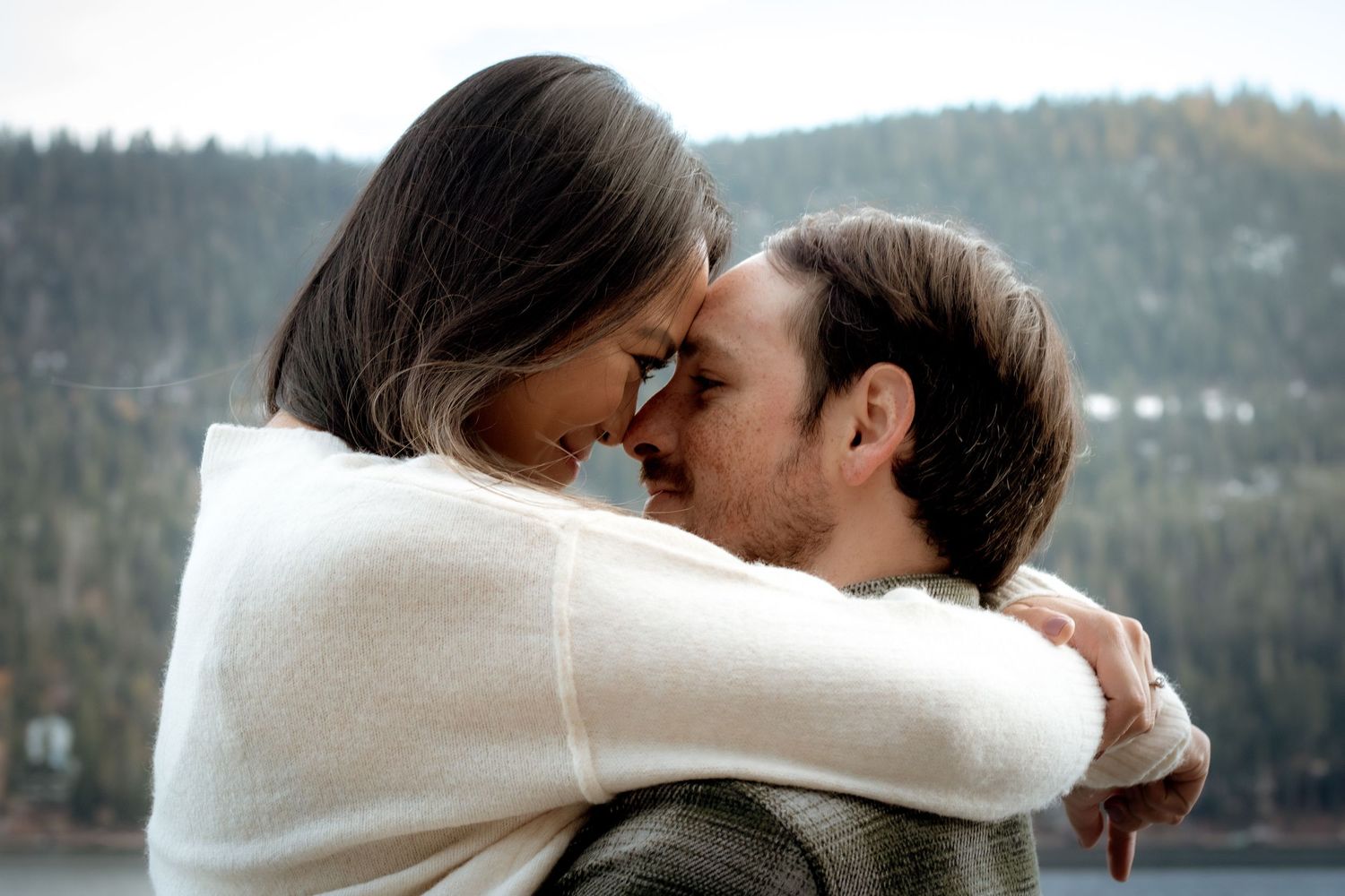 Romantic couple embracing forehead-to-forehead near a misty mountain lake wrapped in a white shawl.