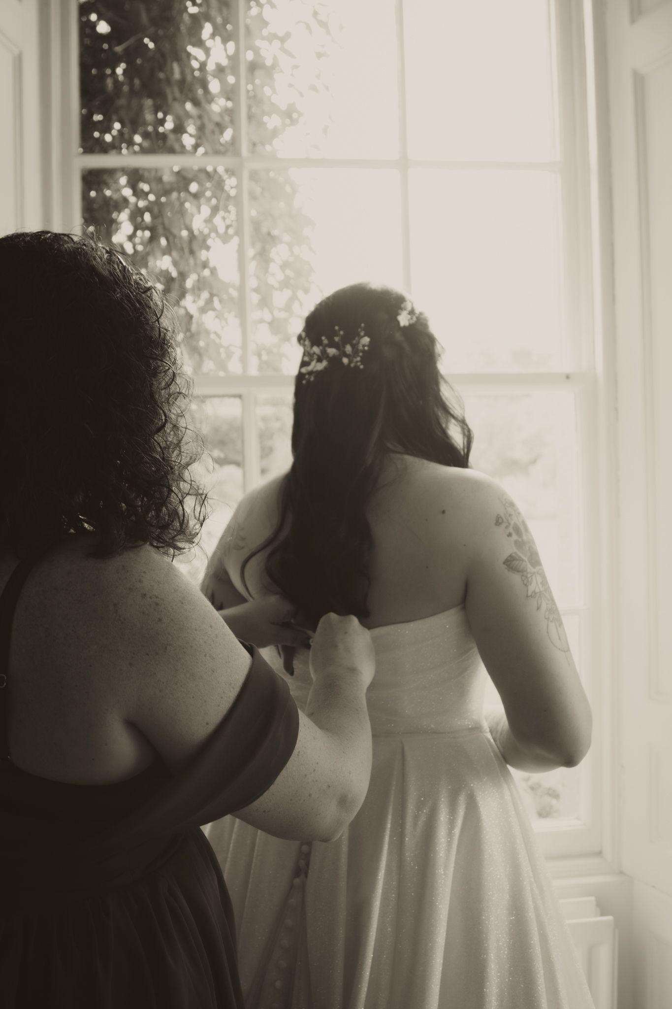 Black and white photo of a bridesmaid helping bride with her dress near a bright window.