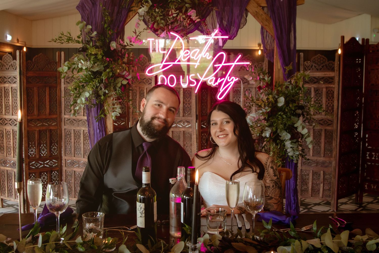 Smiling bride and groom pose at reception table beneath glowing neon wedding sign.