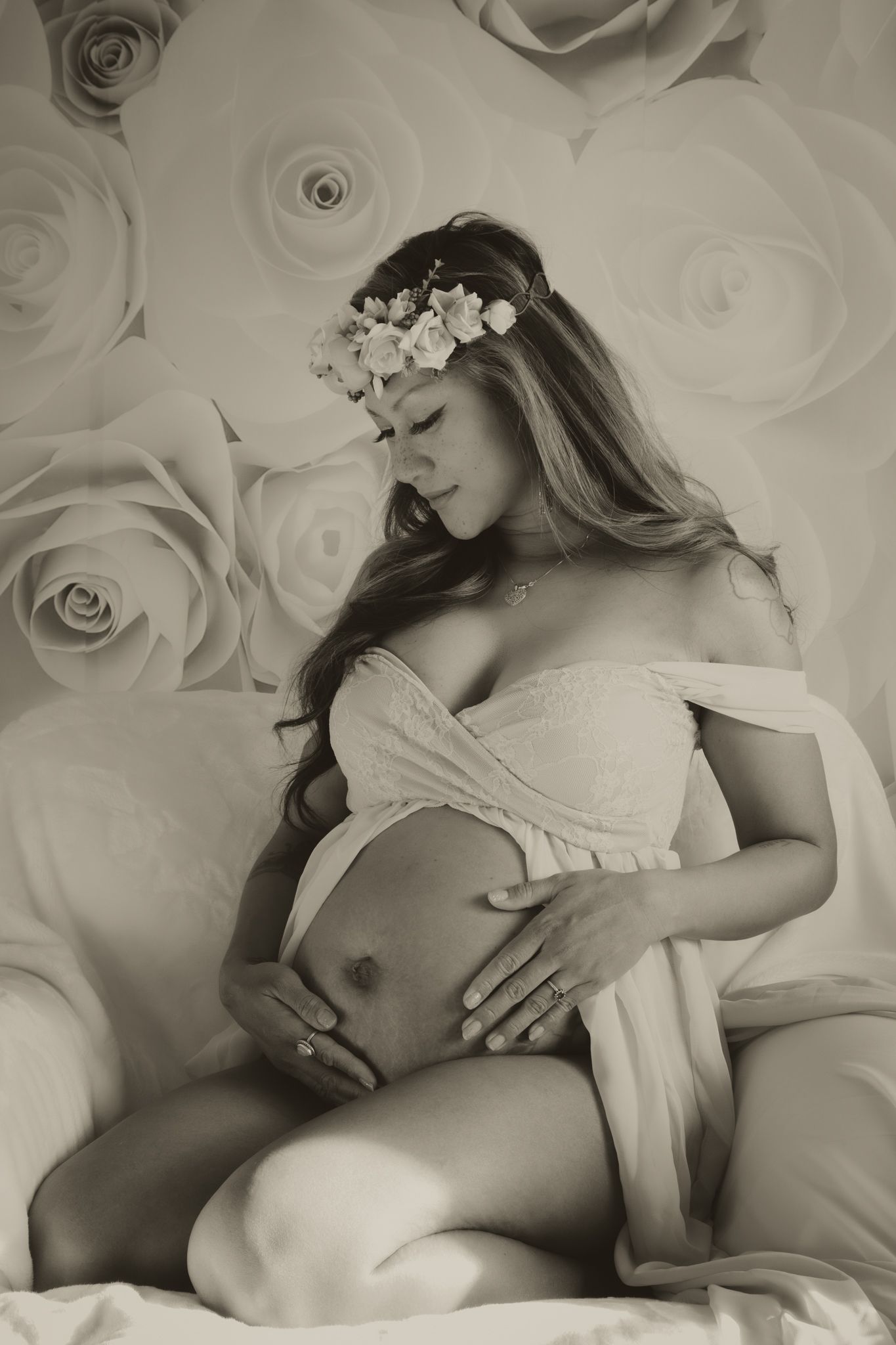 Pregnant woman in white wrap with floral crown poses against rose backdrop in sepia tone.