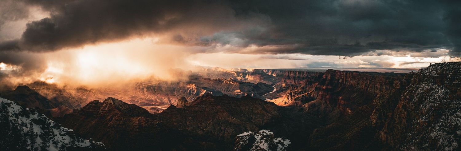 Dramatic panoramic sunset view of the Grand Canyon with stormy clouds and golden light.