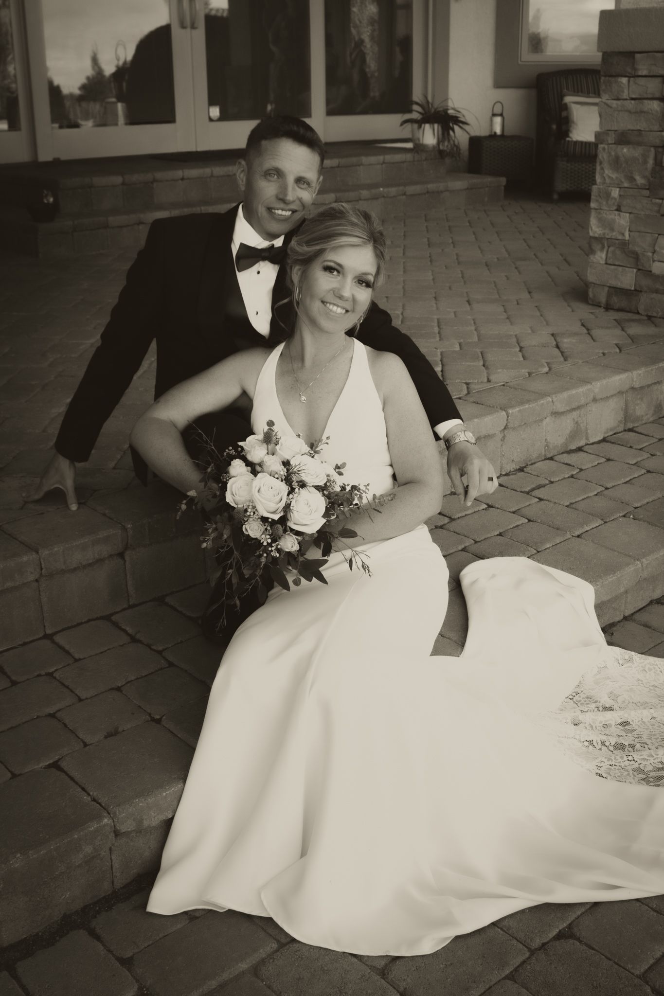 Black and white portrait of smiling bride holding bouquet seated on steps with groom in tuxedo behind her.