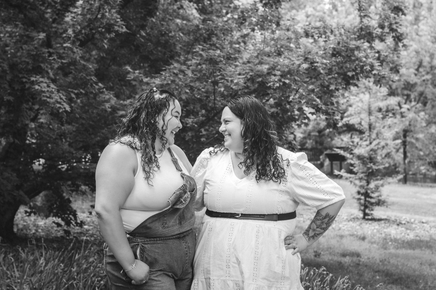 Black and white photo of two women laughing together outdoors among green trees.