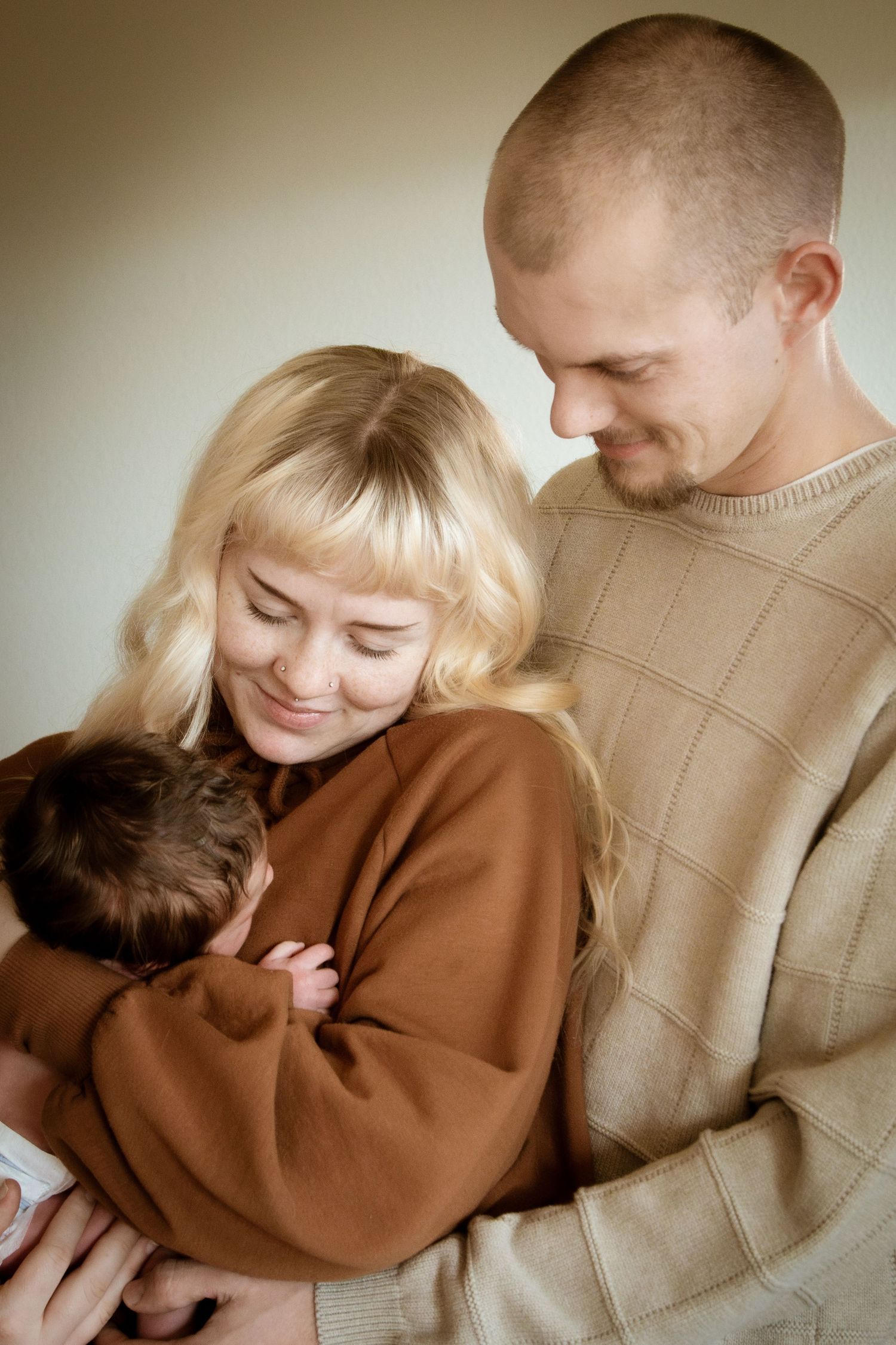 Happy couple smiling and cradling their newborn baby together in a warm, neutral-toned indoor setting.