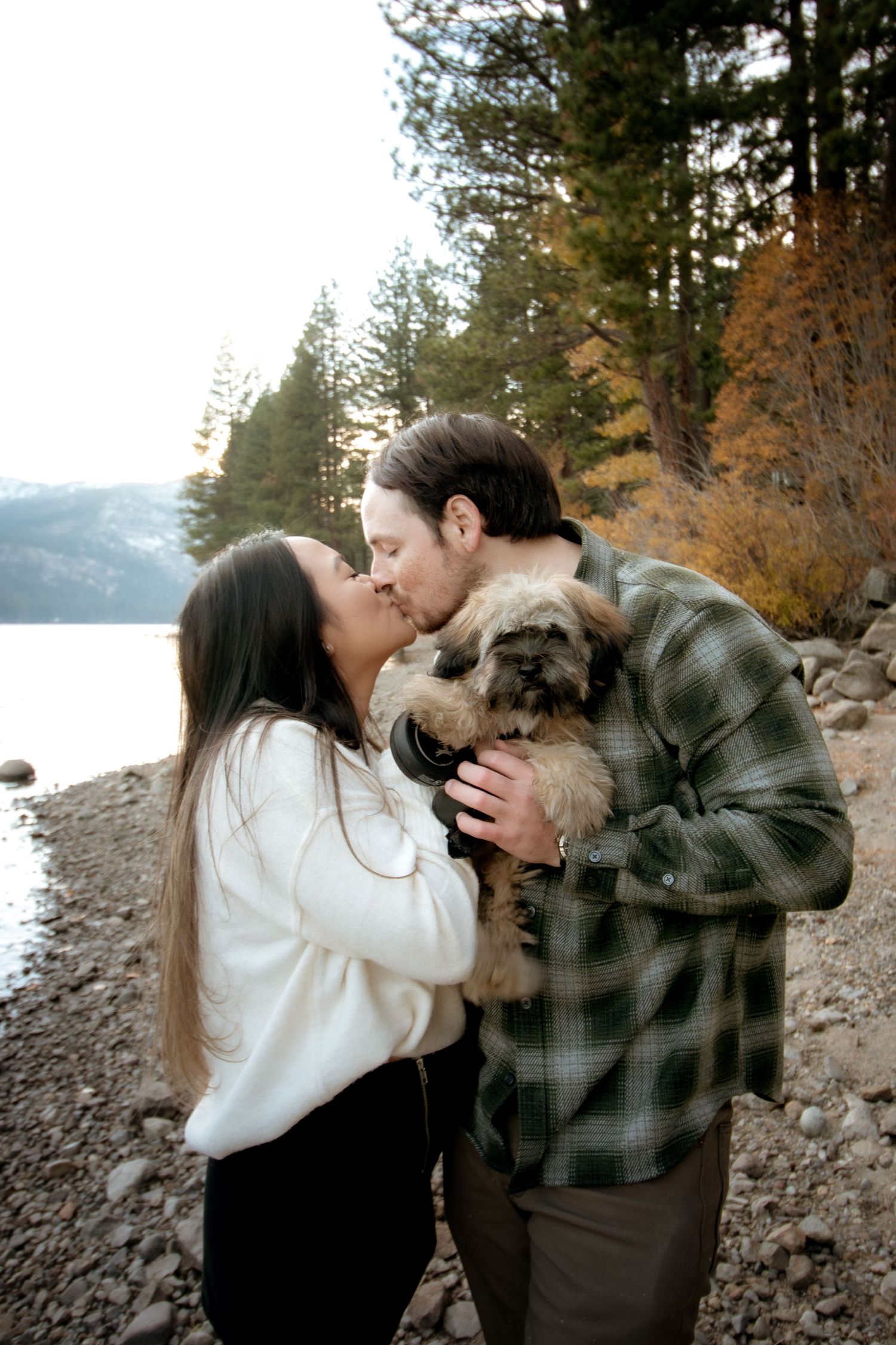 Couple kissing by a lake shore while holding a small fluffy dog, surrounded by autumn trees.
