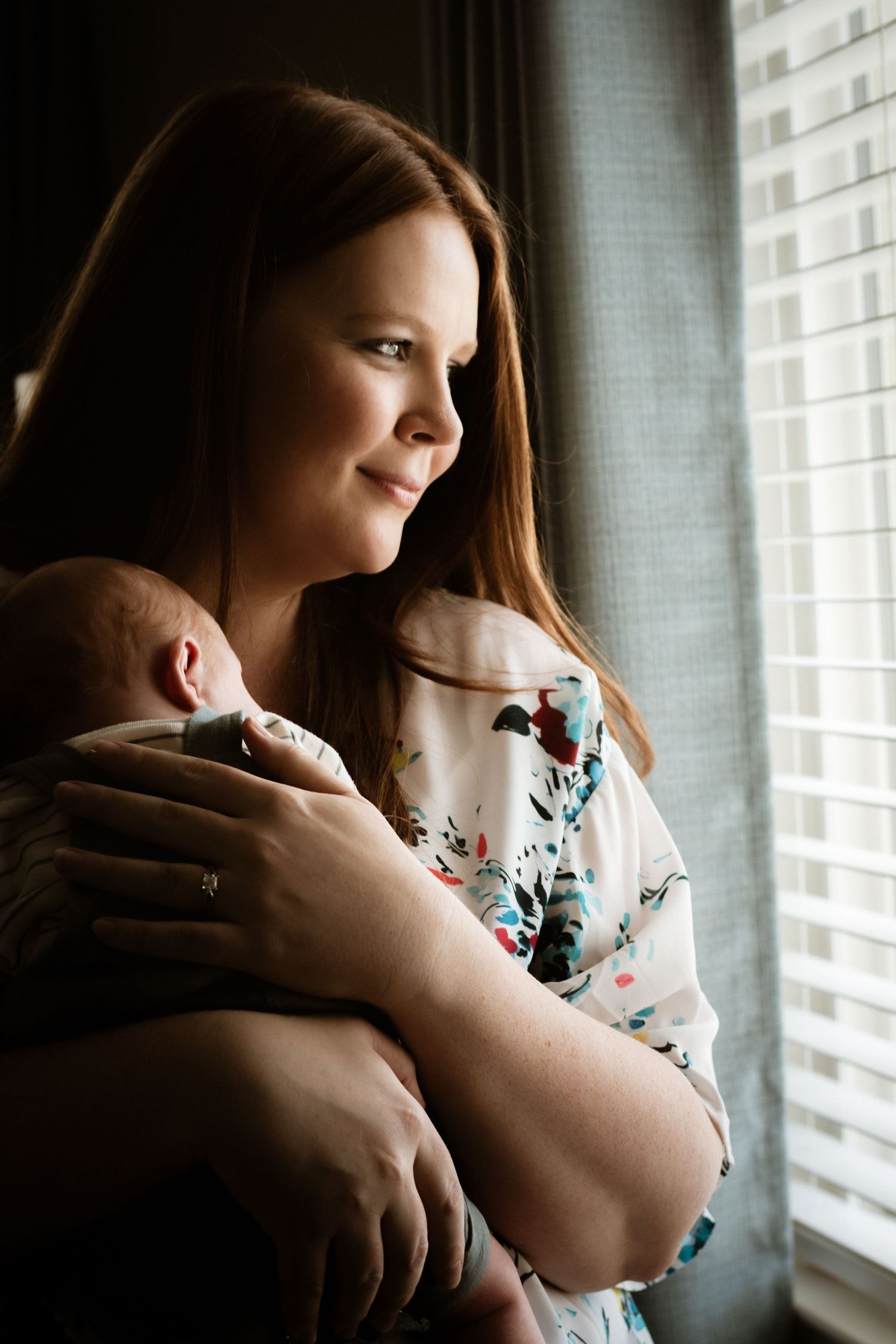 Mother tenderly holds sleeping newborn baby near a sunlit window in floral dress.