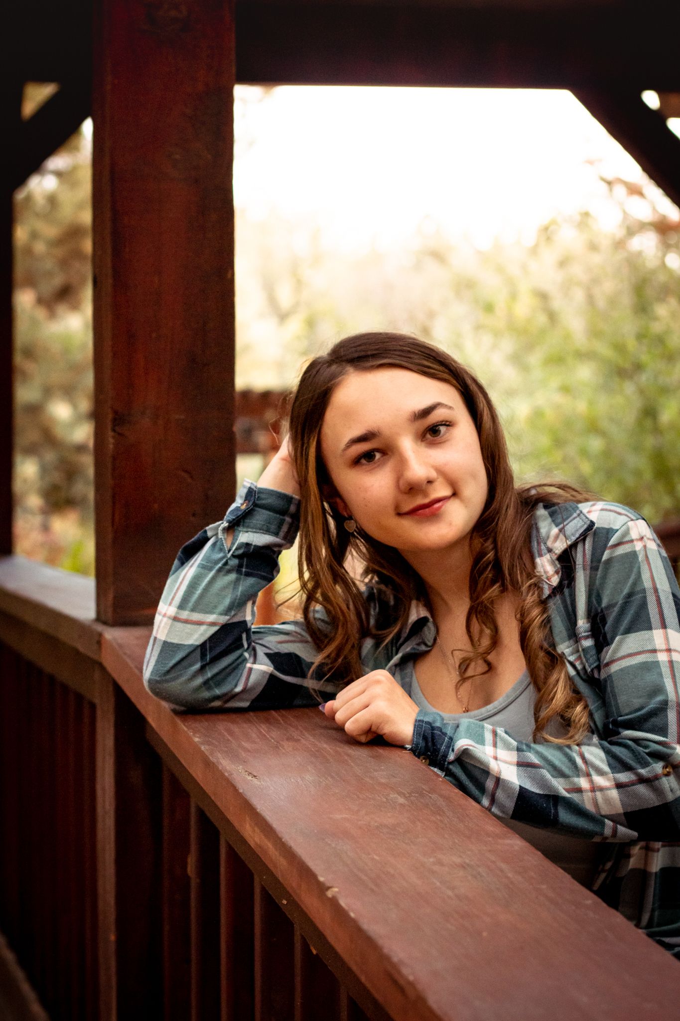 Young woman in plaid flannel shirt leaning on wooden railing, smiling at camera in outdoor setting.