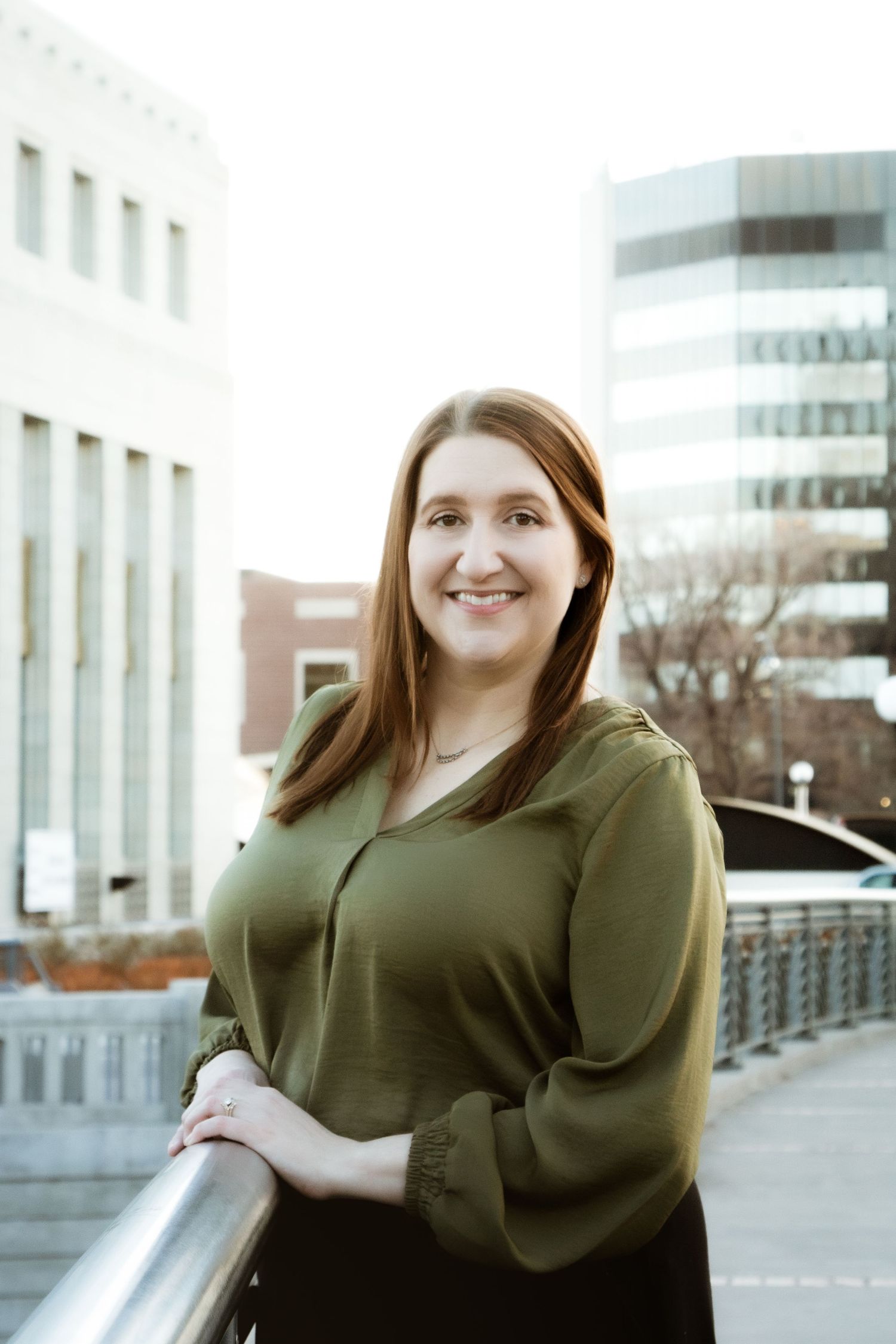Smiling redhead woman in green blouse posing on urban bridge with city buildings in background.