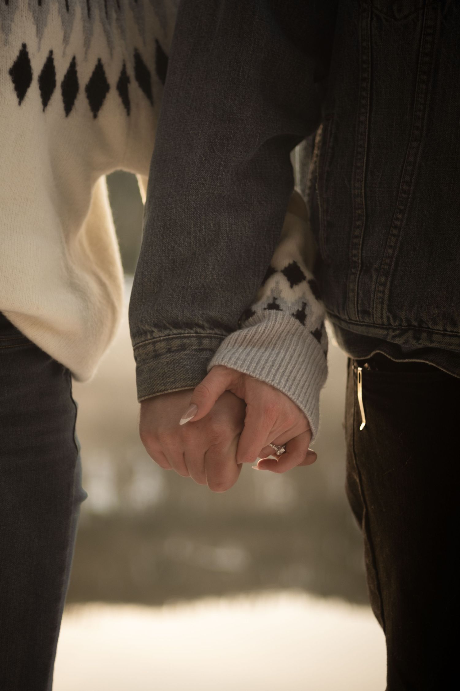Close-up of couple holding hands, woman wearing an engagement ring, dressed in cozy sweaters.