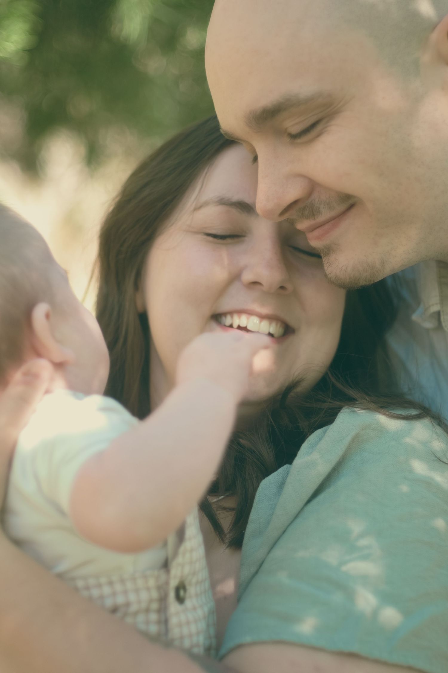 Happy young family with baby sharing a tender moment outdoors, smiling couple touching foreheads with infant.