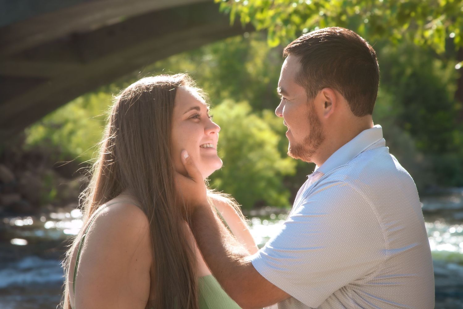 Romantic couple gazing at each other near a river under a bridge in a sunny outdoor engagement photo.