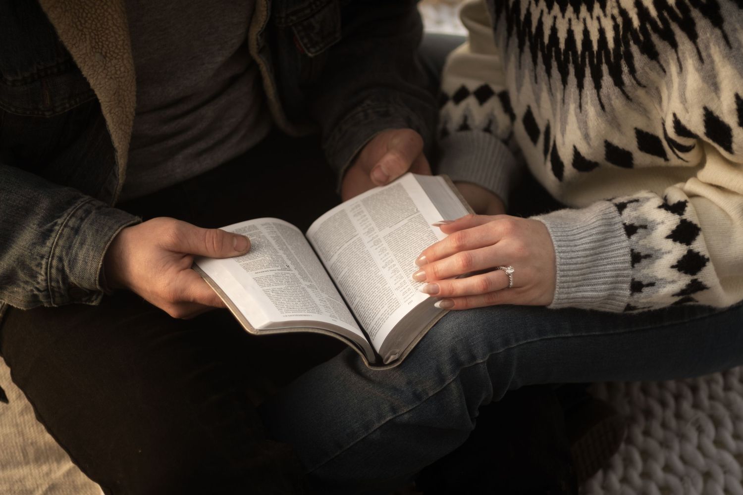 Engaged couple reading Bible together, woman wearing diamond ring, in cozy indoor setting.