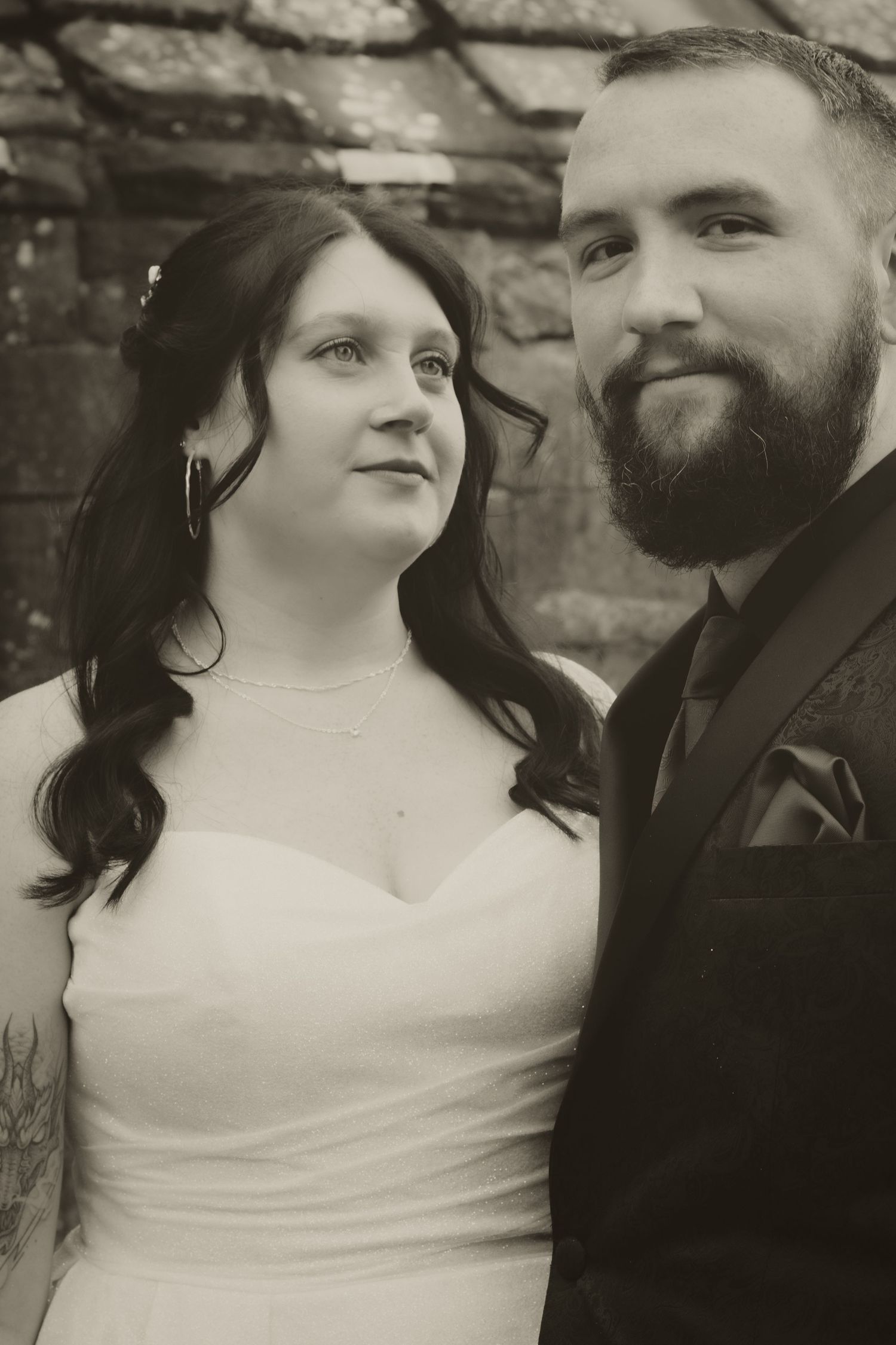 Vintage black and white portrait of bride in strapless dress and bearded groom posing against stone wall.