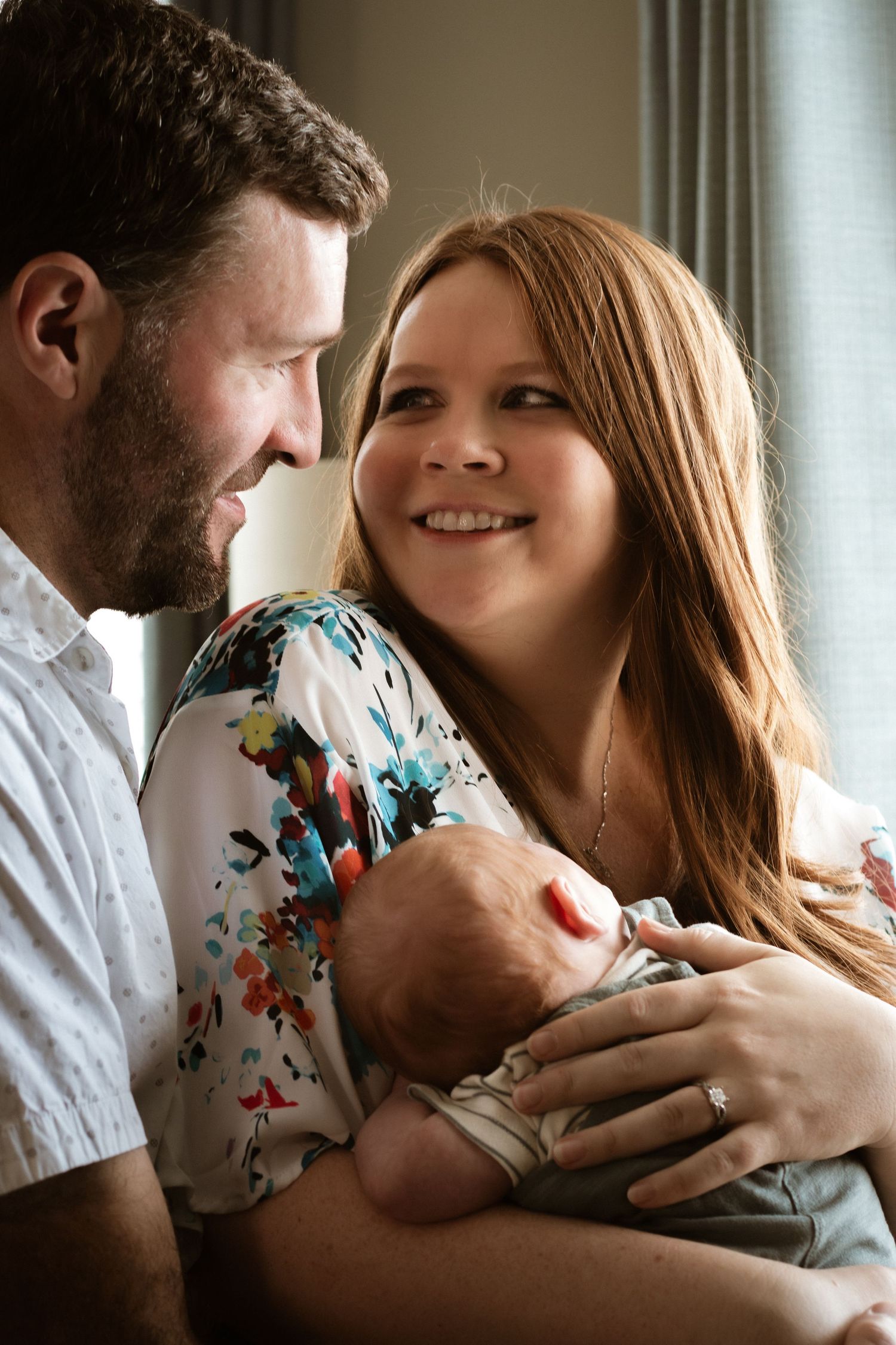 Smiling new parents holding their newborn baby together near a window in a warm lifestyle family portrait.