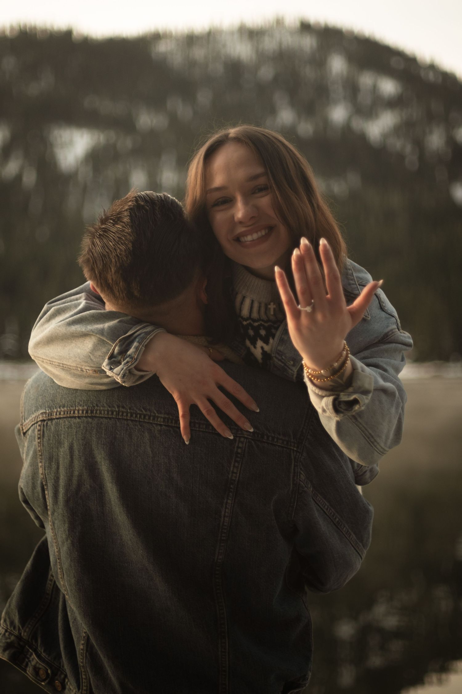 Joyful woman showing engagement ring while embracing her partner outdoors with snowy mountains behind.
