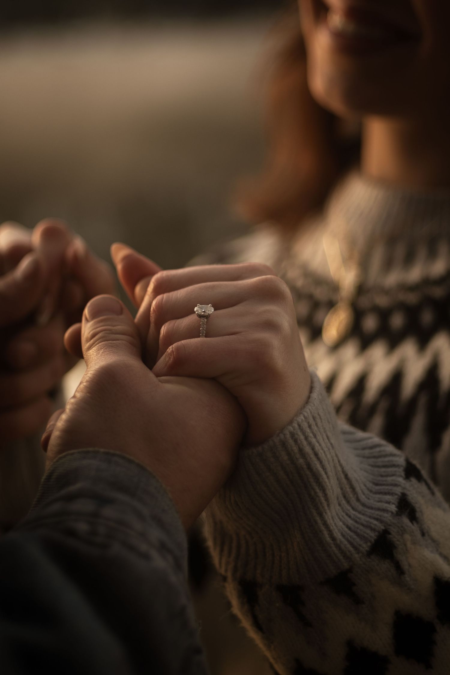 Close-up of a couple holding hands showing an engagement ring, wearing cozy sweaters in warm golden light.