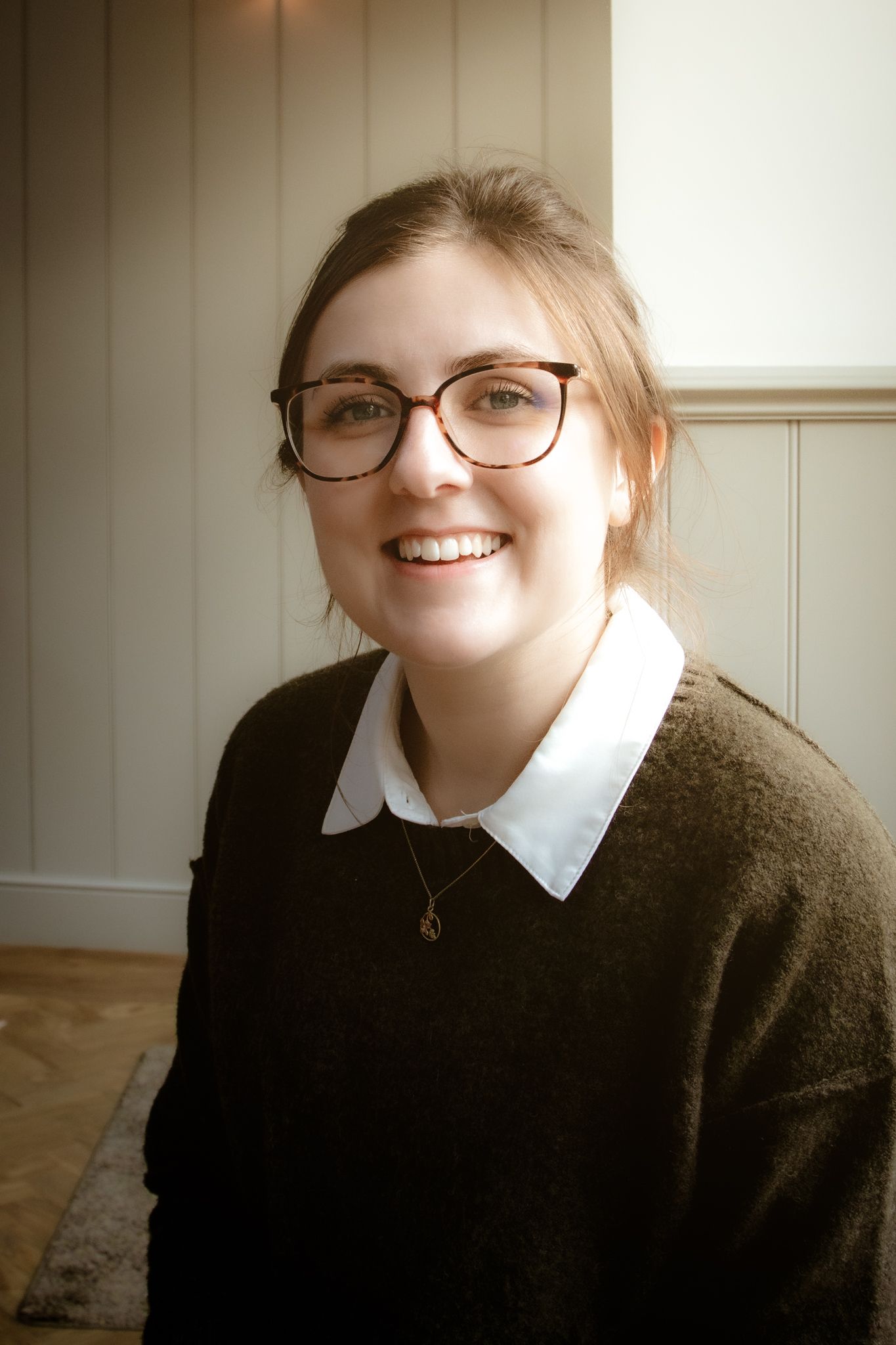 Cheerful young woman wearing tortoiseshell glasses and dark sweater with white collar smiling indoors.
