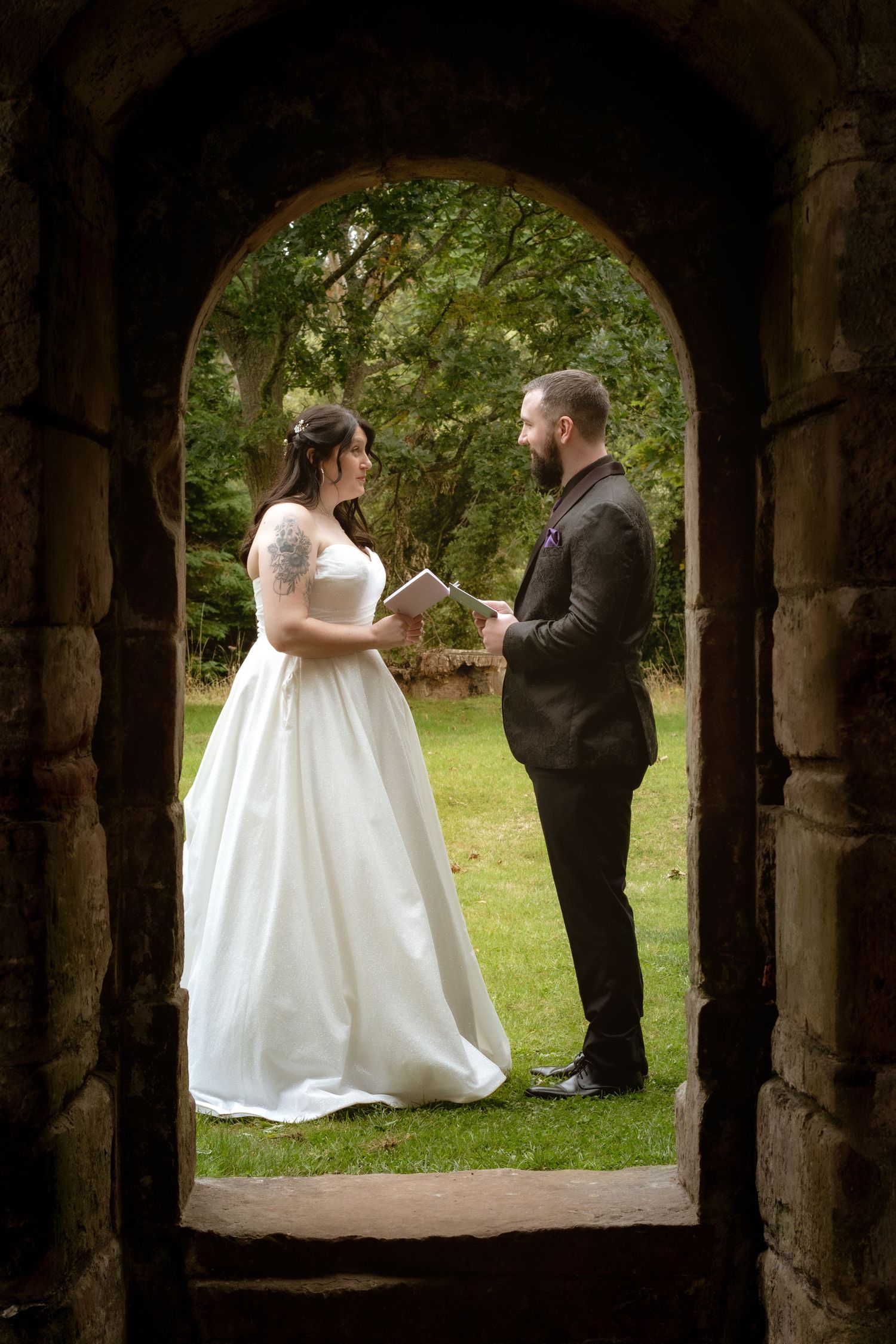 Bride and groom exchange vows framed by a romantic stone archway surrounded by lush green garden foliage.