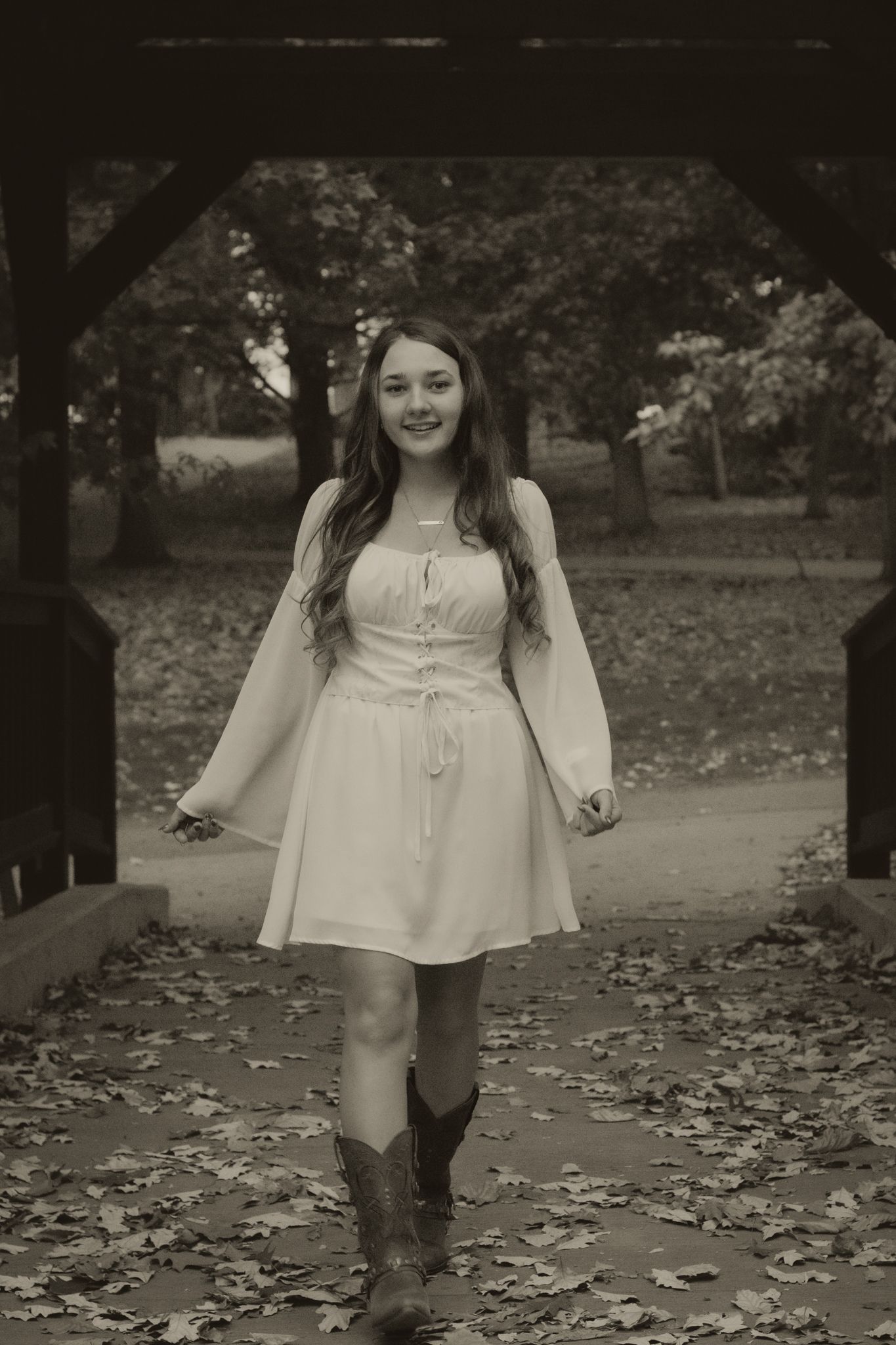 Young woman in white dress and cowboy boots walks under wooden pergola in sepia-toned autumn park photo.