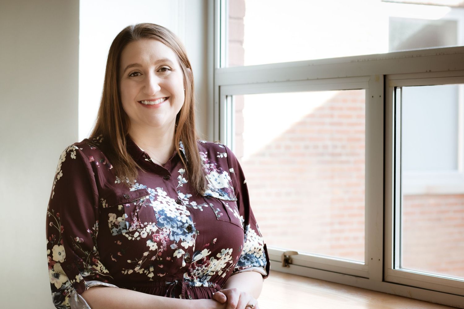Smiling woman in floral burgundy blouse poses by bright window in professional indoor portrait.