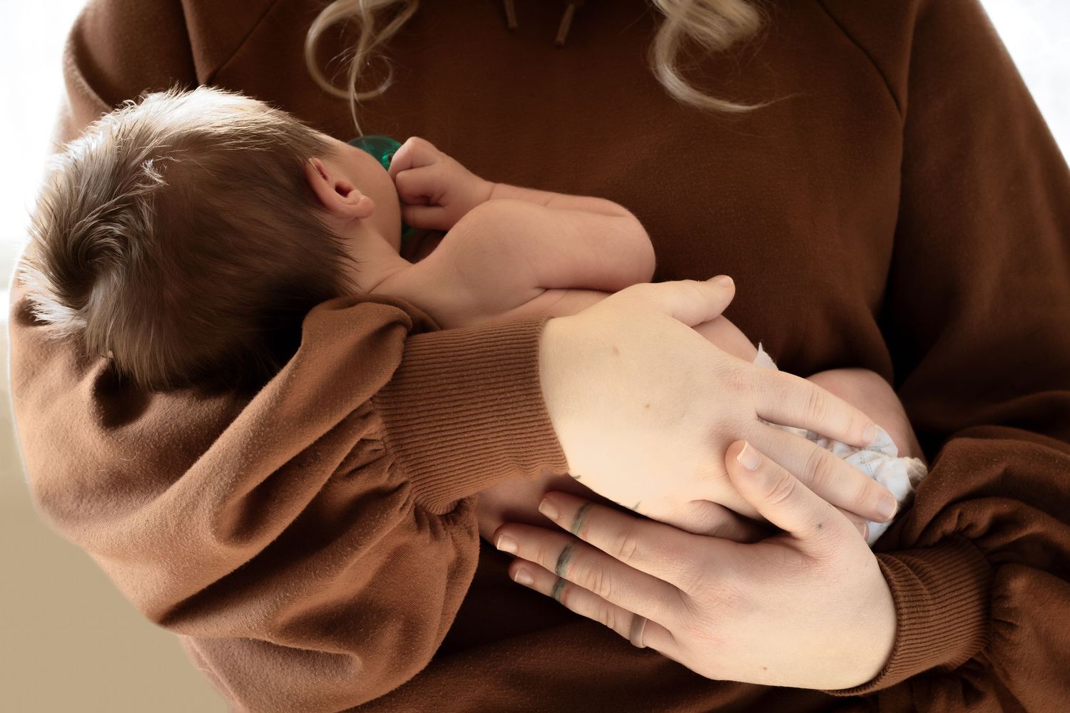 Mother in brown hoodie gently cradling sleeping newborn baby in her arms.