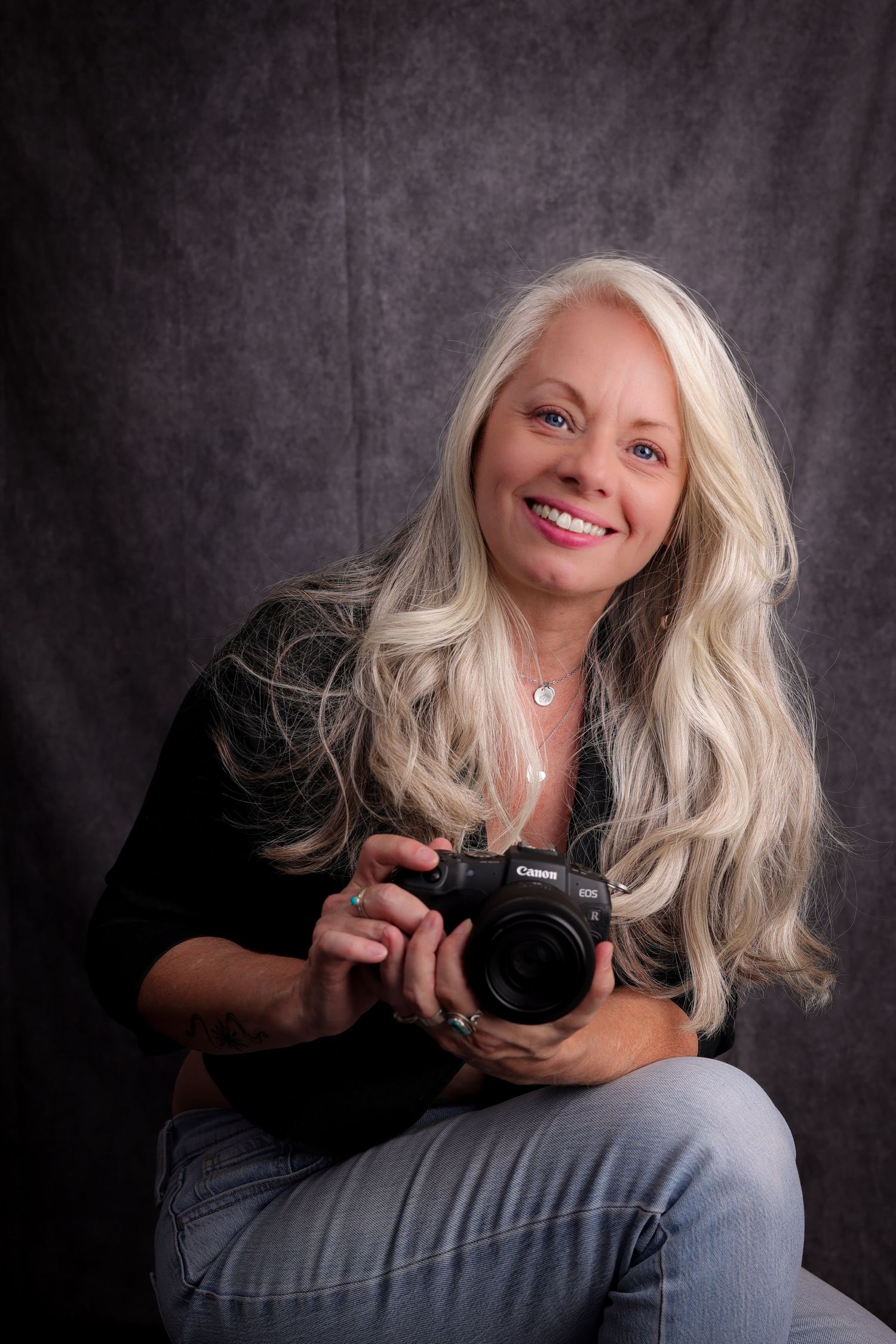 Professional photographer with blonde hair wearing black top and jeans poses with camera against dark background.
