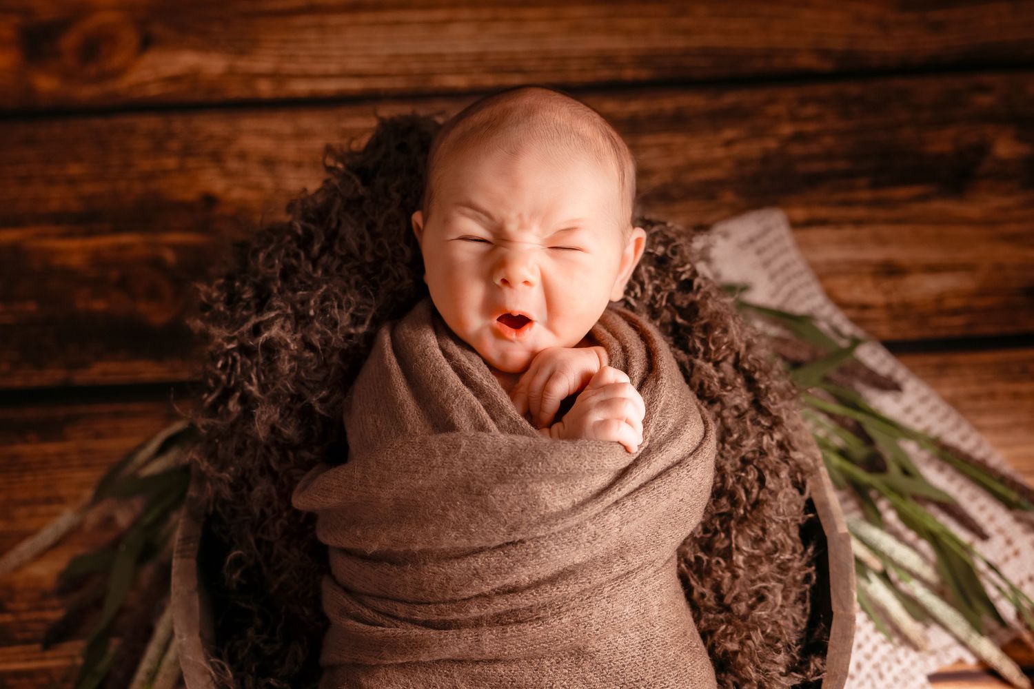 Newborn baby wrapped in brown blanket lying on rustic wooden backdrop during professional photography session.