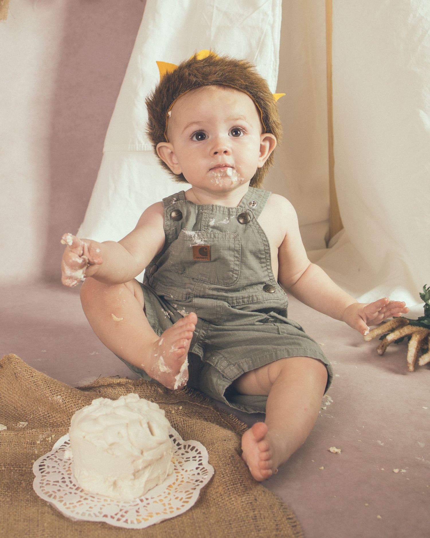 A baby in green overalls sits during a first birthday cake smash photo session.
