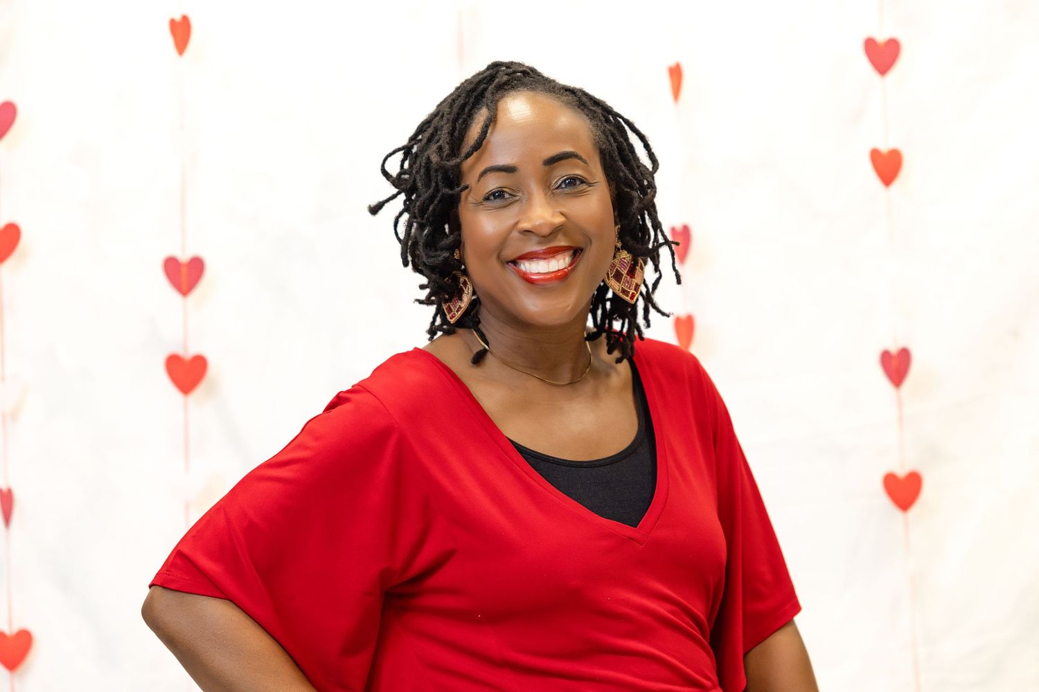A person wearing a red dress smiles warmly against a white background with small pink heart decorations.