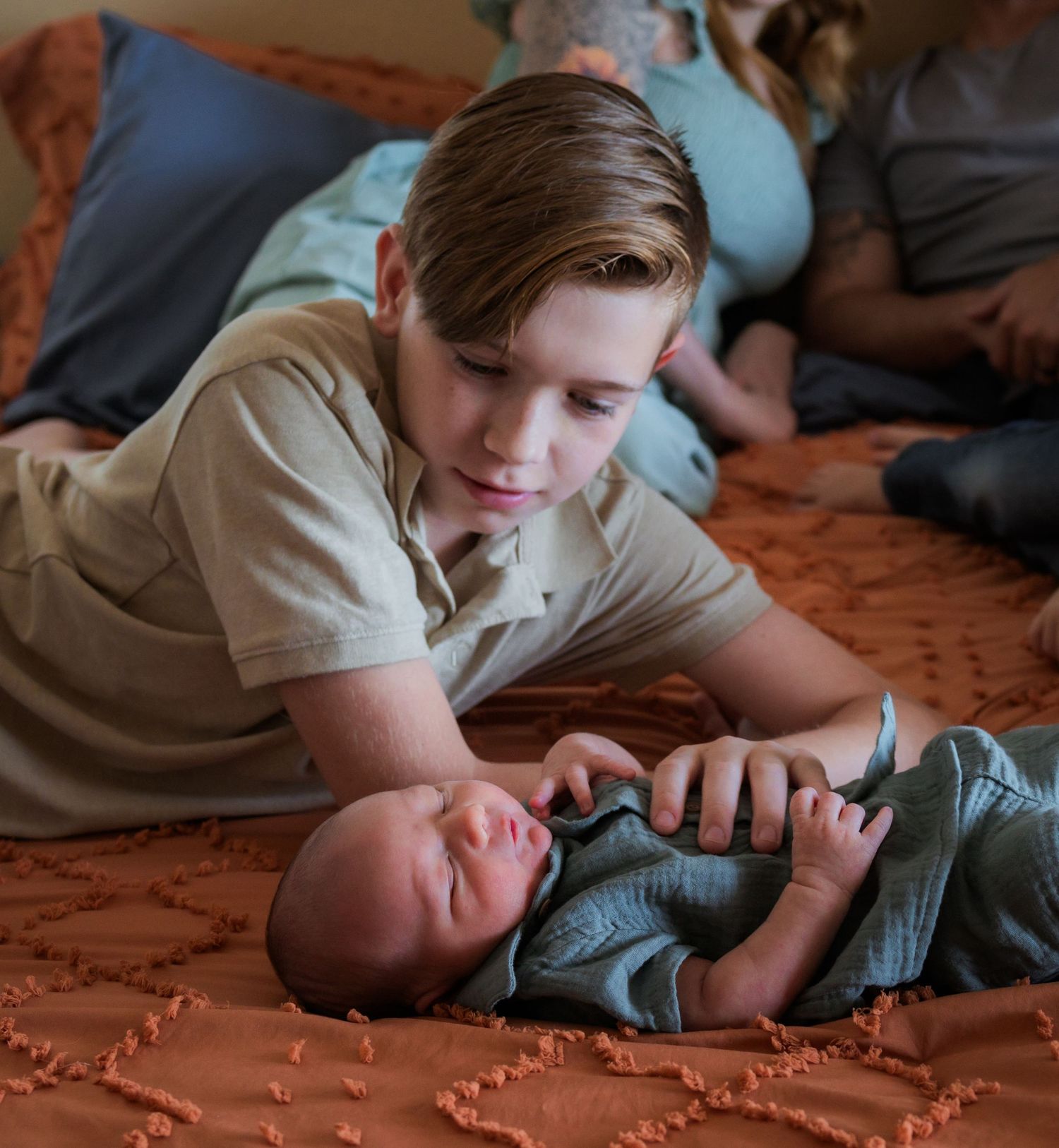 Older brother with newborn baby lying on a patterned bed comforter in a relaxed pose.