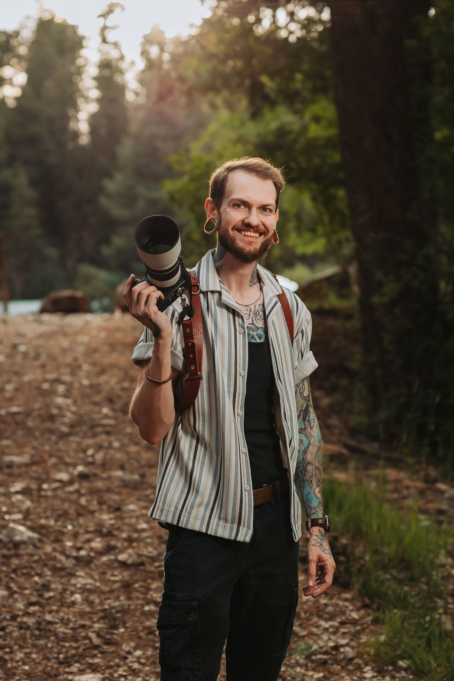 Smiling tattooed photographer holding a camera with telephoto lens in a sunlit forest setting.