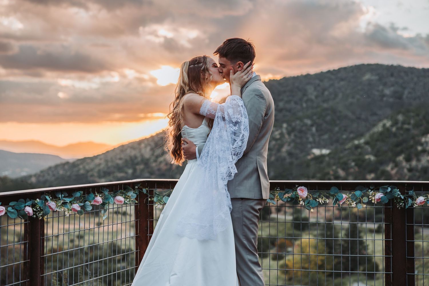 Romantic couple embraces on a mountain overlook during a dramatic sunset with city views in the background.