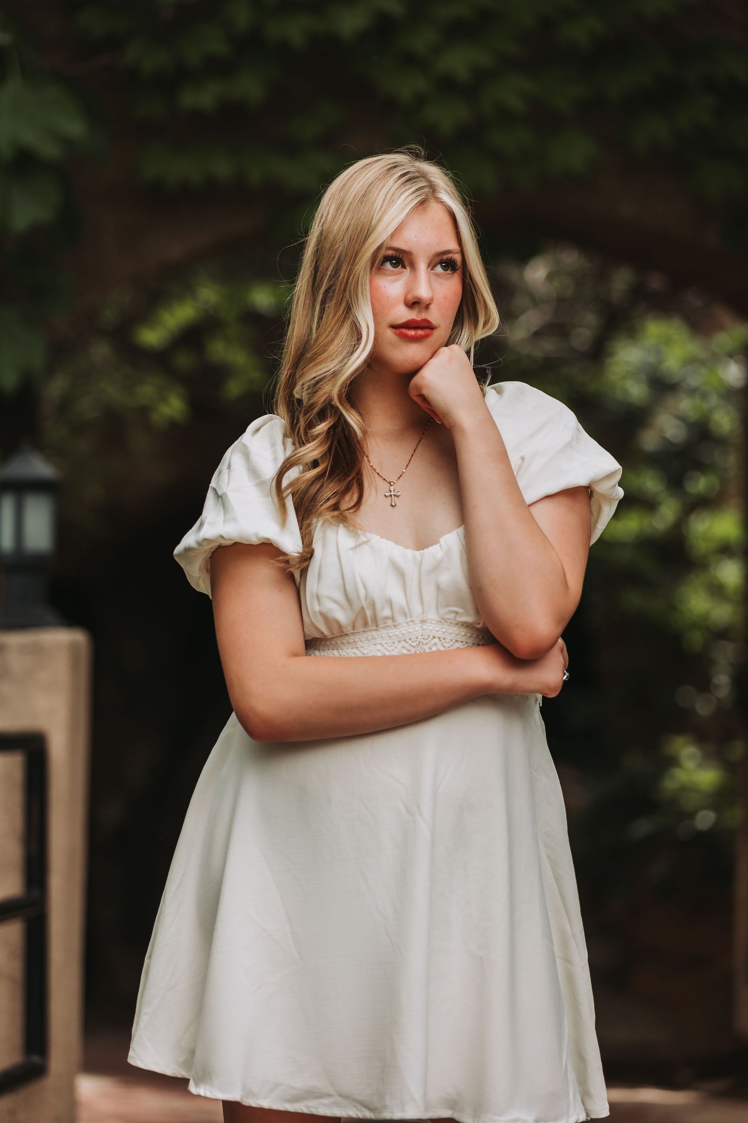 Woman in white dress poses thoughtfully in outdoor garden setting with natural lighting and greenery background.