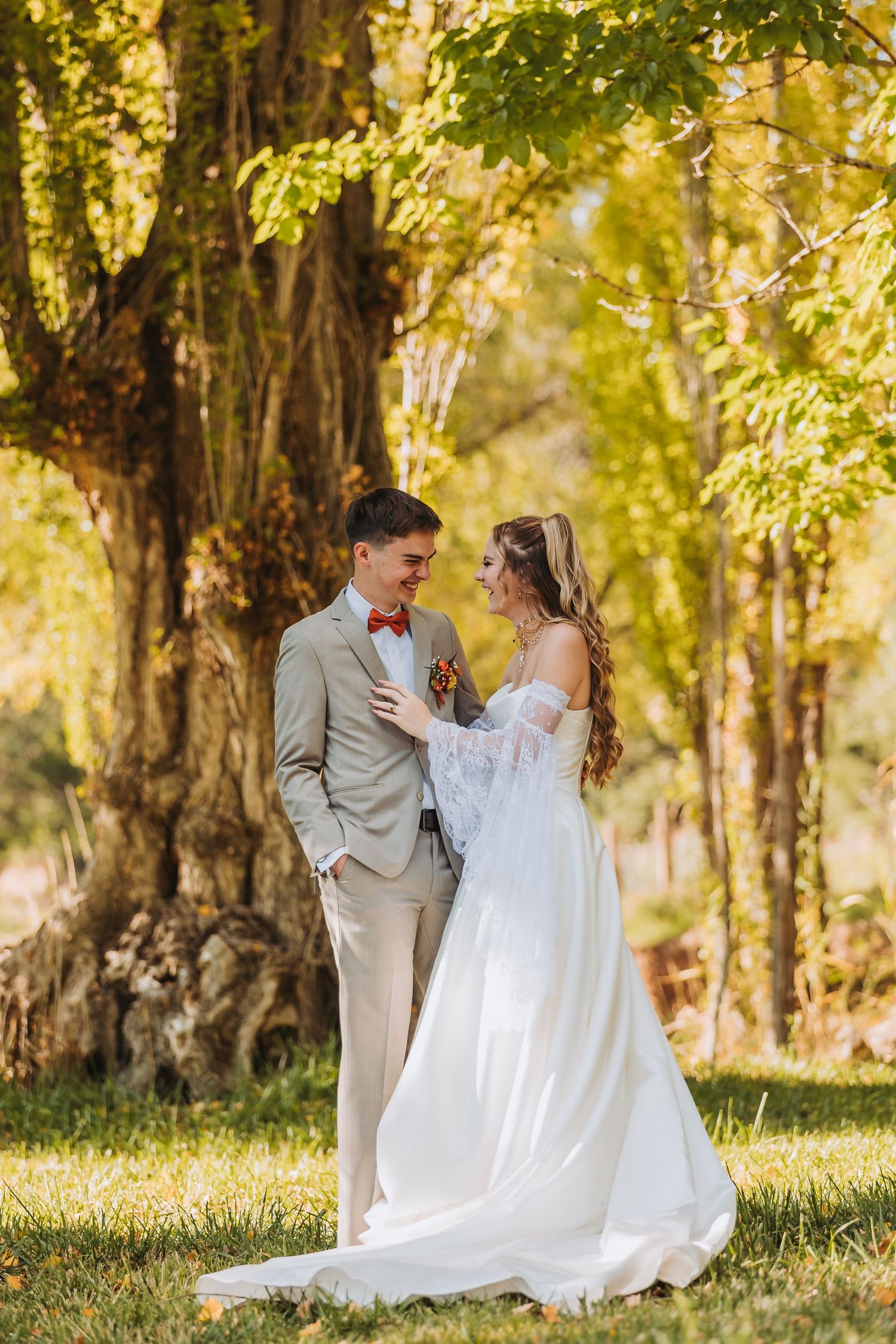 A couple in wedding attire share an intimate moment under sunlit trees in an autumn outdoor setting.