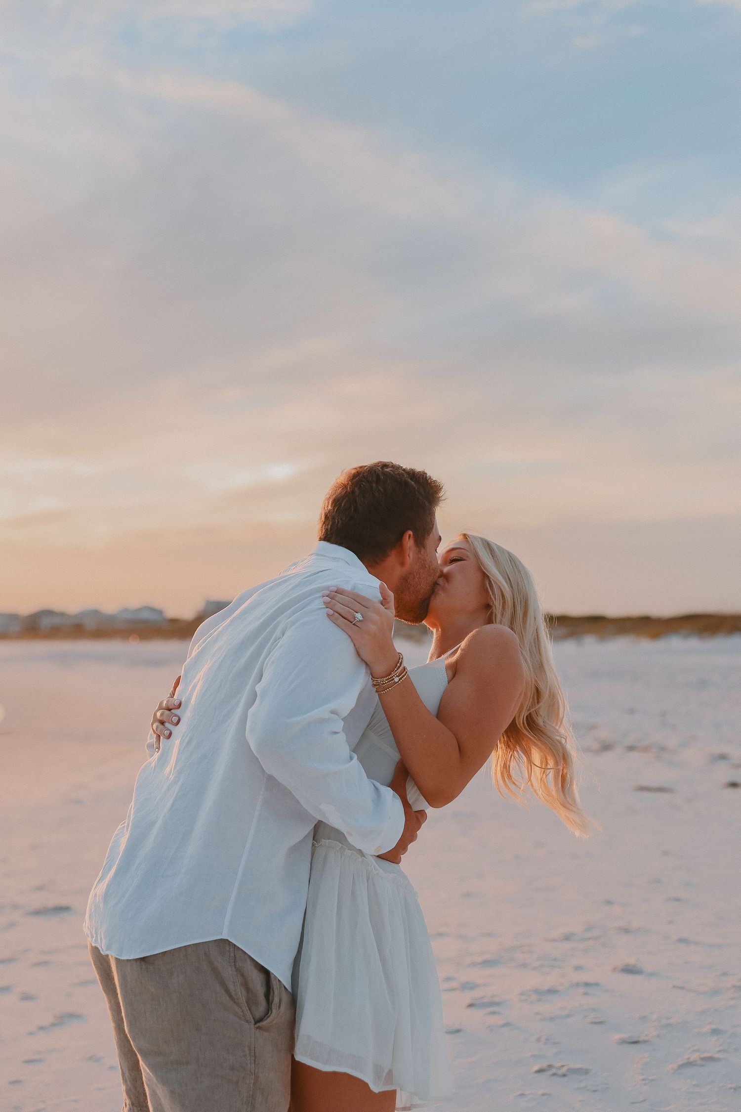 Couple sharing a romantic kiss on a sandy beach at sunset, woman in white dress with long blonde hair.
