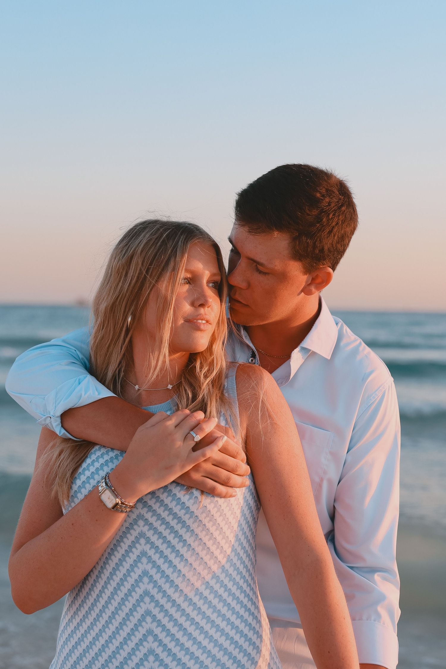 Couple embracing on beach at sunset, man kissing woman's cheek, wearing white shirt and blue dress.