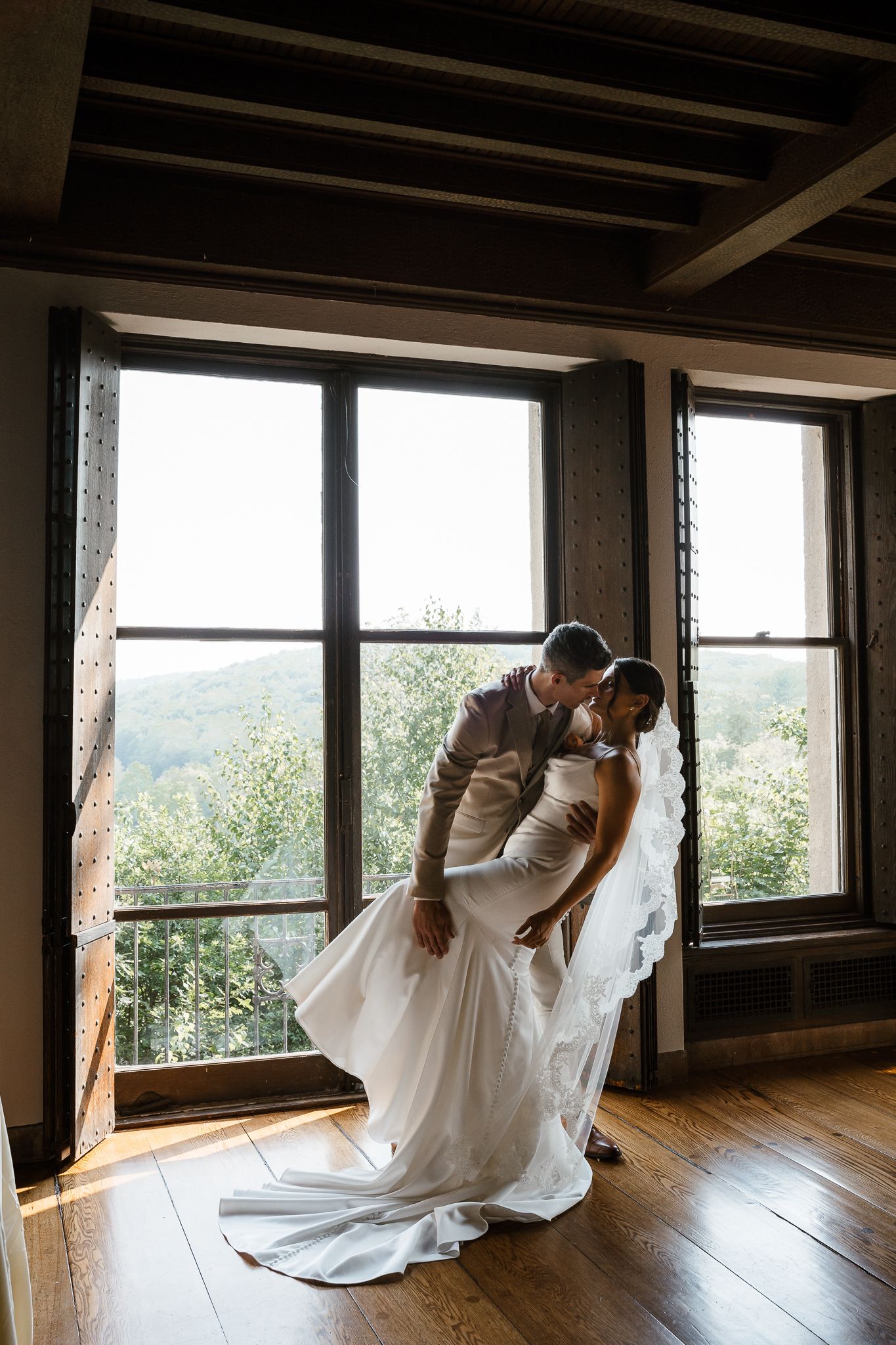Bride and groom share a romantic dip kiss by large windows with scenic mountain views in a rustic wood-floored venue.