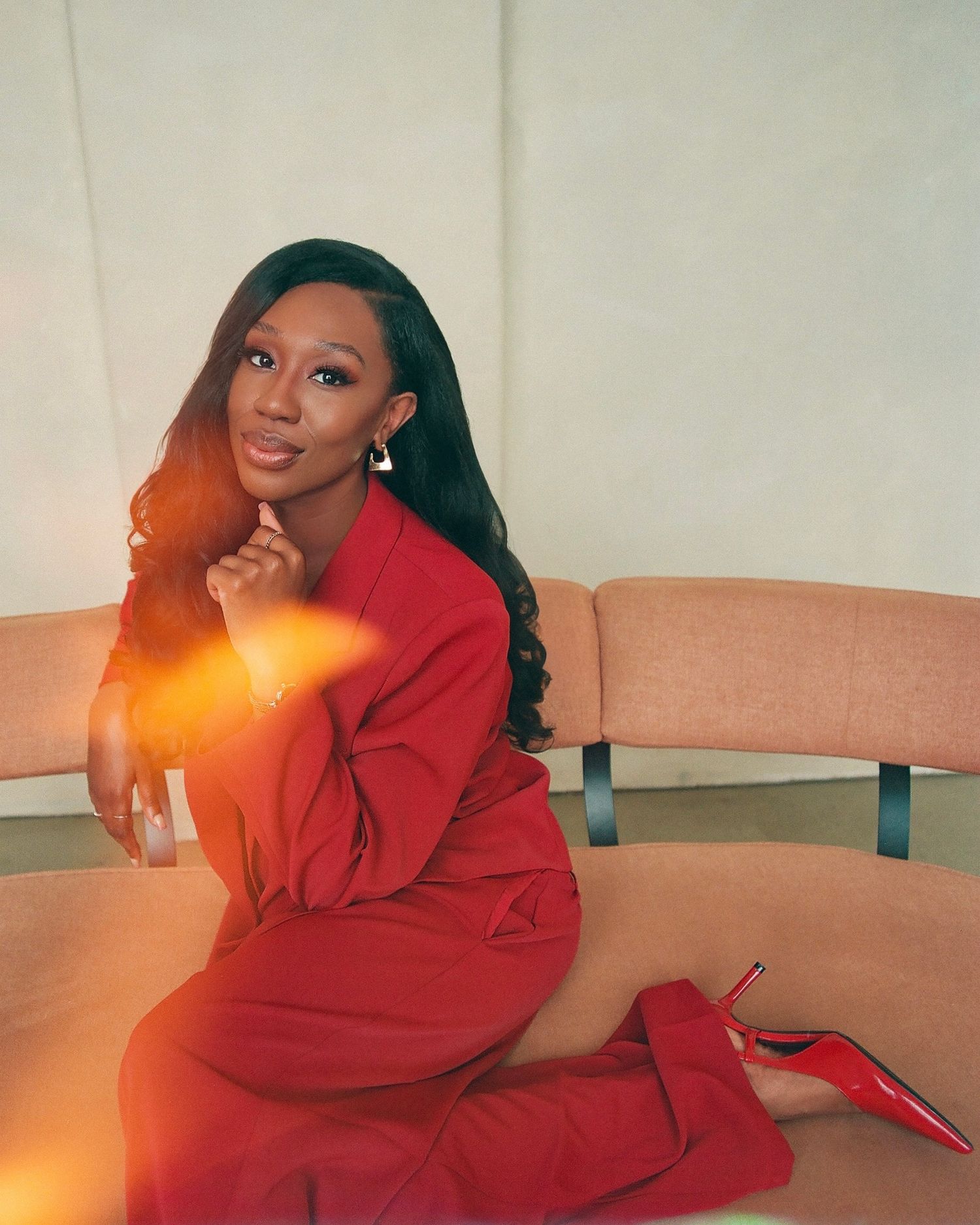 A woman in a red suit and heels poses elegantly on a beige sofa in a modern interior setting.