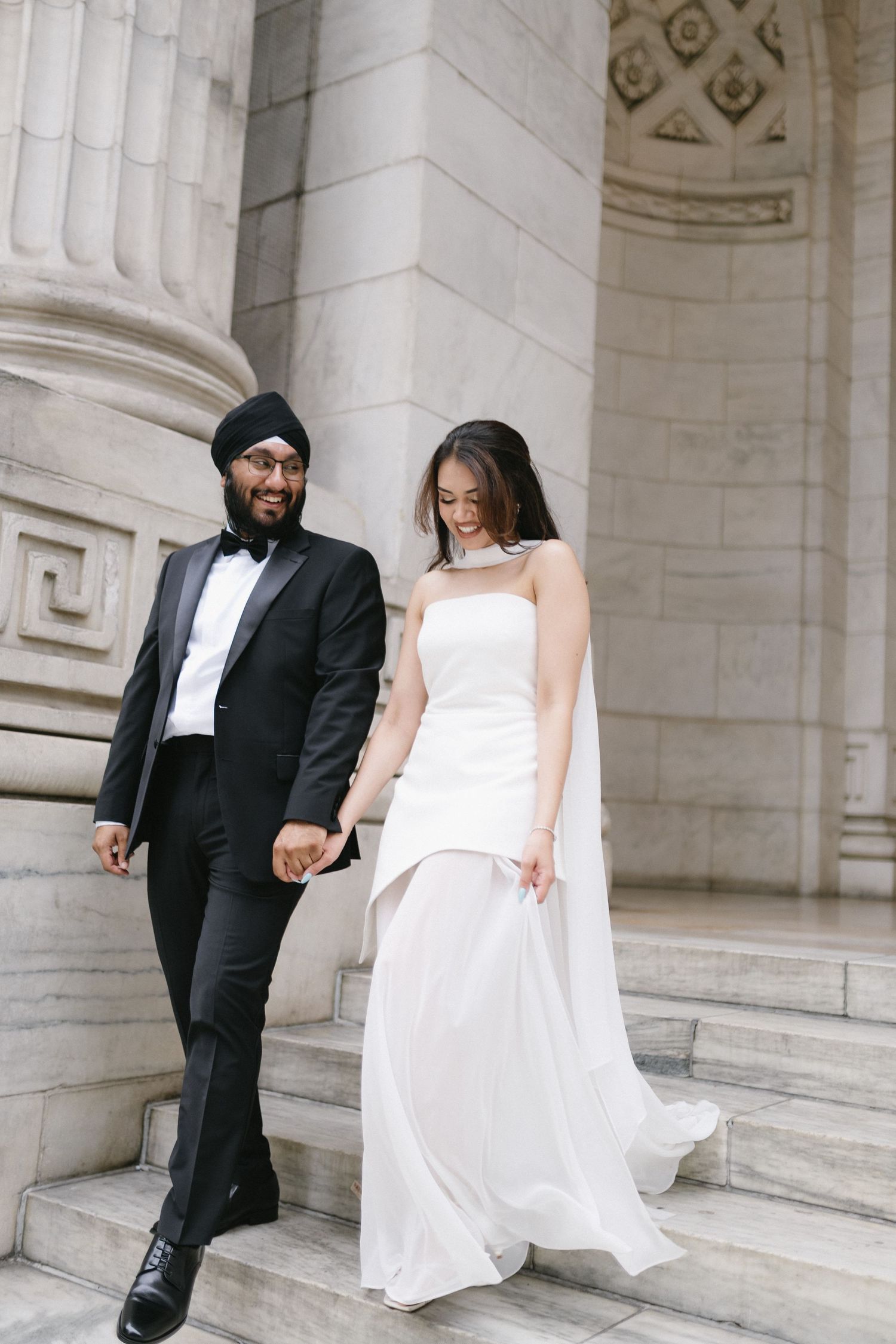 A Sikh groom in a black tuxedo and turban holds hands with his bride in a white gown on marble steps.