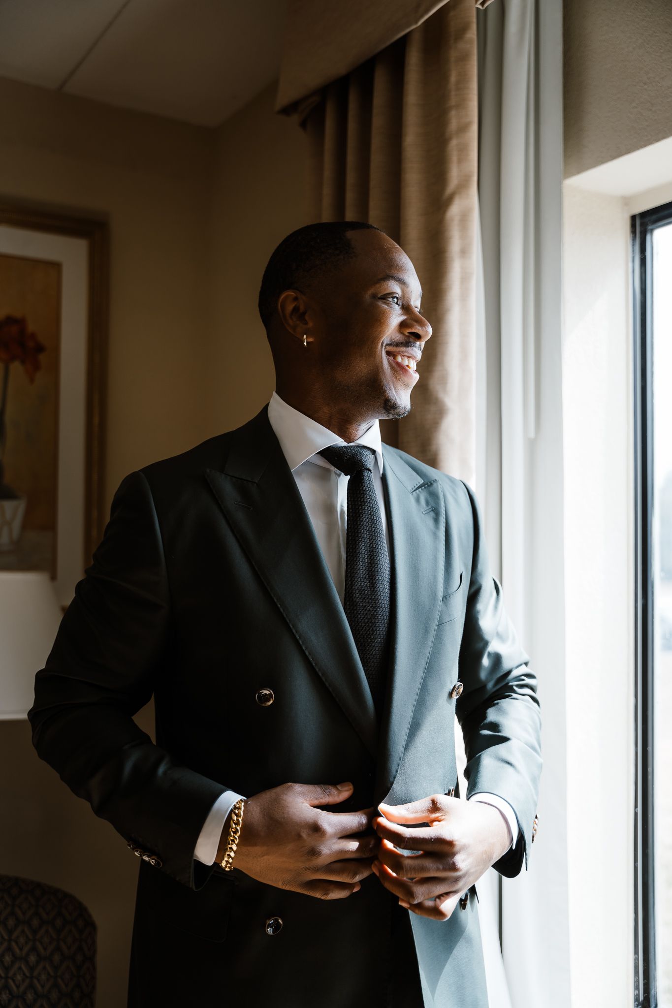A well-dressed person in a dark suit stands by a window, smiling warmly while adjusting their jacket.