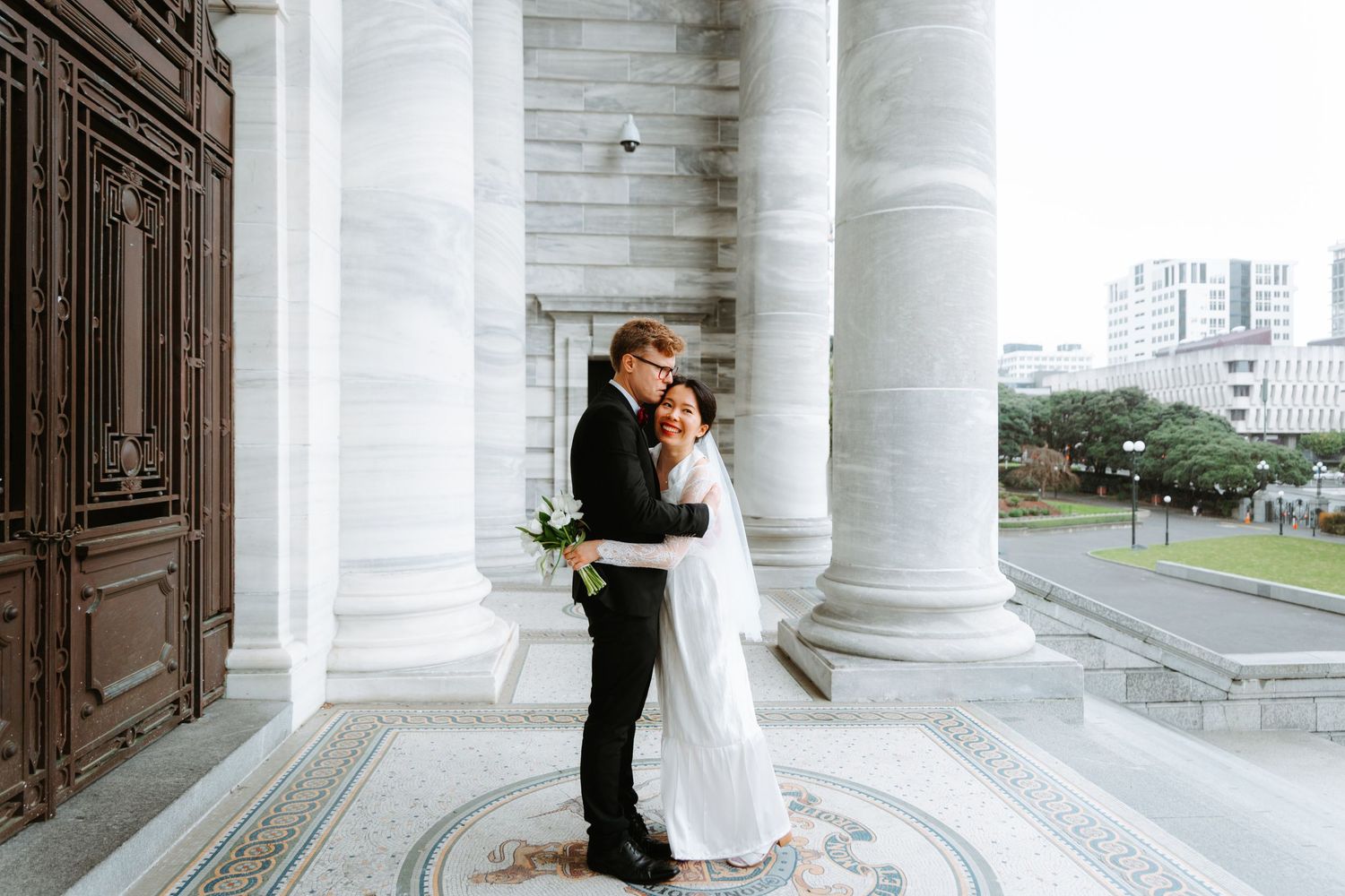 A couple embraces in their wedding attire between elegant white marble columns of a classical building.